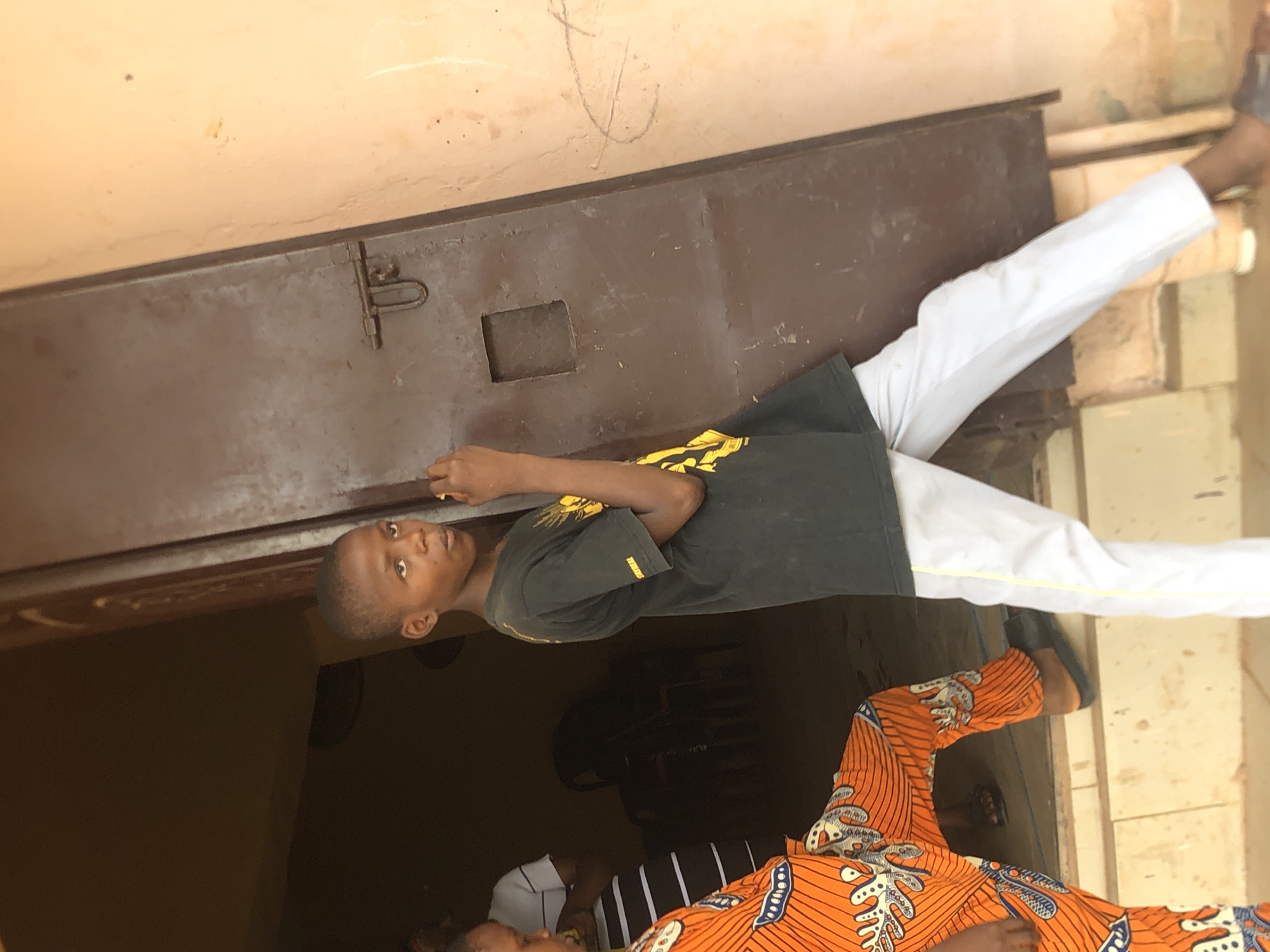 A young boy is standing partially in front of a large, dark, rectangular metal structure, possibly a gate or door, with white trousers. He is wearing a black t-shirt with a yellow design. The top of the structure is attached to a light brown wall. In the foreground, another person's legs are visible, clad in orange patterned fabric with blue details. The scene is likely outdoors, possibly in or near Nkwelle Ezunaka, Nigeria. There are other partial figures visible, including what looks like a person in a white shirt with black stripes, lying on the ground. The lighting is somewhat uneven, with shadows and bright spots.