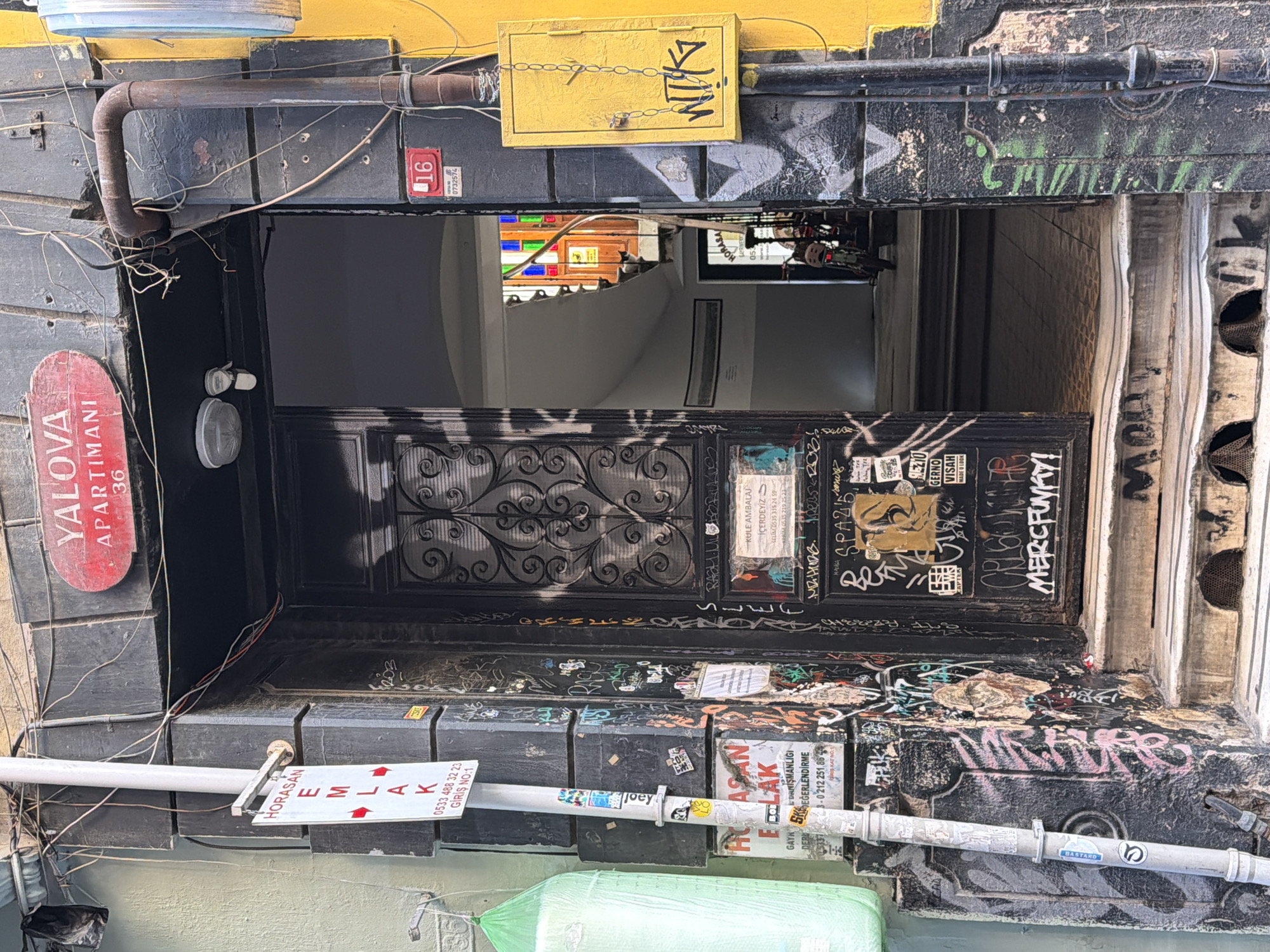 The image shows the entrance to an apartment building in Beyoğlu, Turkey. The building facade is dark gray and covered in graffiti, with a bright yellow electrical box mounted on the right side. A red sign above the entrance reads "YALOVA APARTMANI 36". The doorway is open, revealing a dimly lit staircase with stained glass windows at the top. A motorcycle is parked on the stairs. A white pole with several signs is visible to the left of the entrance, also covered in graffiti. The steps leading up to the entrance are weathered and also bear graffiti. The overall impression is one of a well-worn urban environment with a strong artistic, albeit informal, presence.