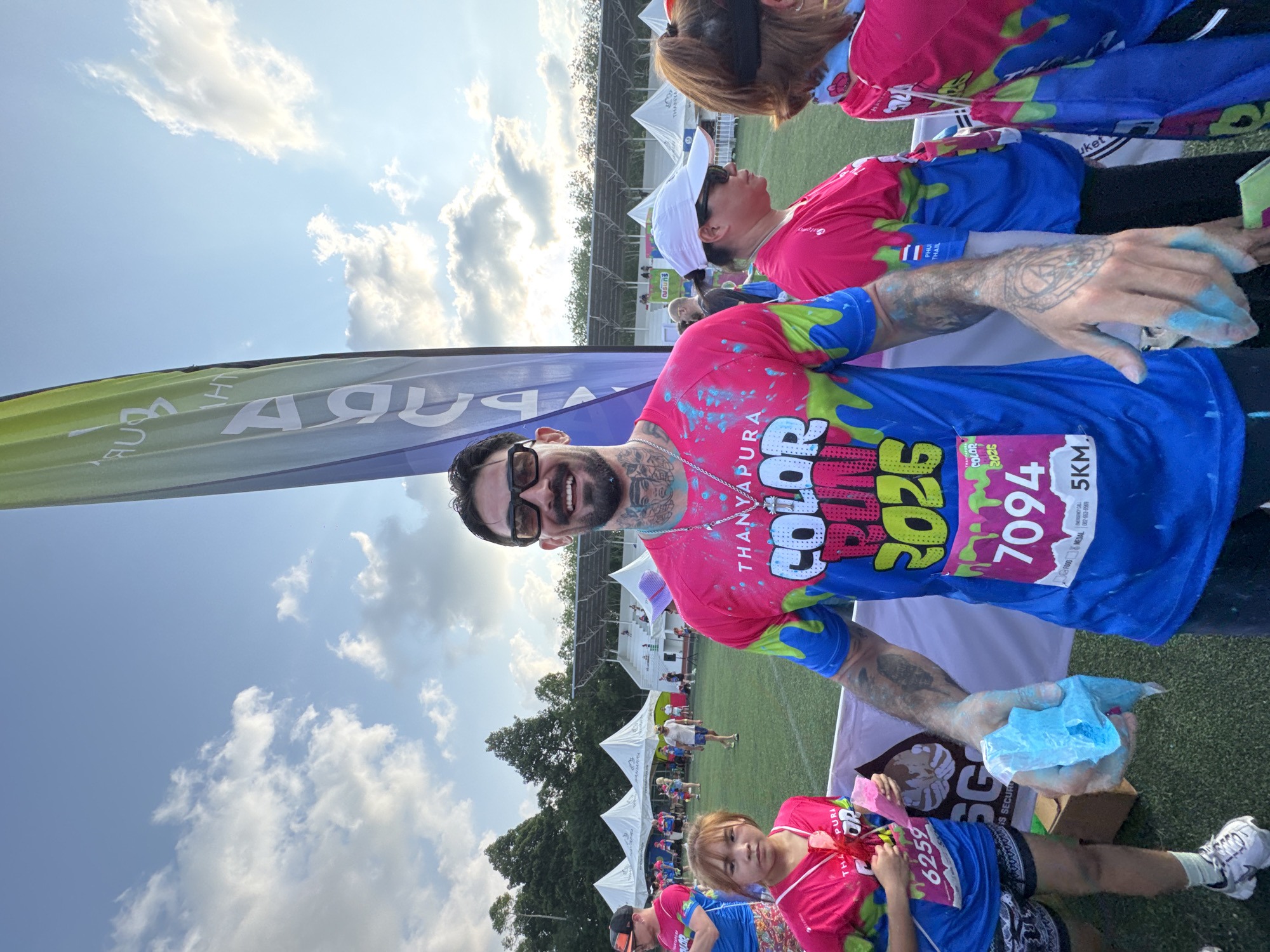 The image captures a scene in Thep Krasatti, Thailand, featuring a man and a girl participating in the Thanyapura Color Run 2026. The man, wearing glasses and adorned with tattoos, is holding a bag of blue powder, indicative of the color run event. He is smiling at the camera. Both the man and the girl are wearing matching t-shirts. The girl appears to be holding a bag of pink powder. Their shirts are splattered with various colors.
In the background, other participants can be seen, along with white tents and green fields. A banner bearing the word "THANYAPURA" is visible. The sky is bright with scattered clouds. Race bibs on the shirts display the numbers "7094" and "6259." The event appears to be held outdoors on a grassy field.