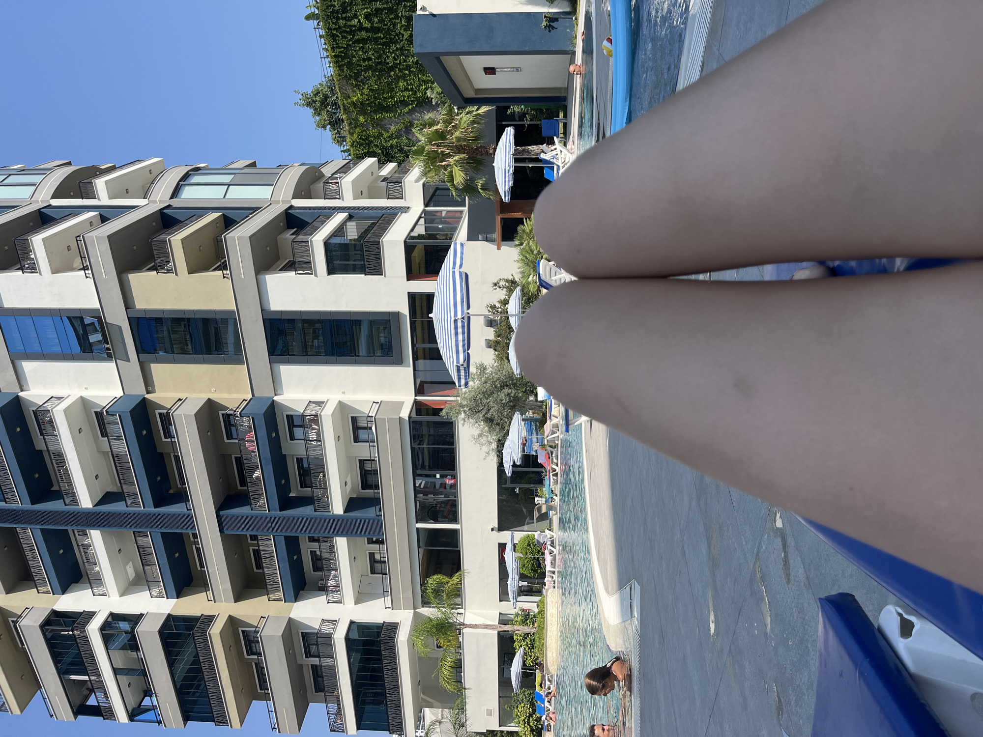 The image shows a close-up of a person's legs stretched out, likely on a lounge chair, in the foreground. Beyond the legs, a swimming pool is visible with people in and around it. Blue and white striped umbrellas are set up around the pool area, and lounge chairs are scattered on the deck. In the background, a modern, multi-story building with balconies and blue-tinted windows dominates the scene. Green vegetation is visible on the right side of the building. The sky is clear and blue, indicating a sunny day. The context of Alanya, Turkey, suggests this is likely a resort or hotel with a pool and apartments.