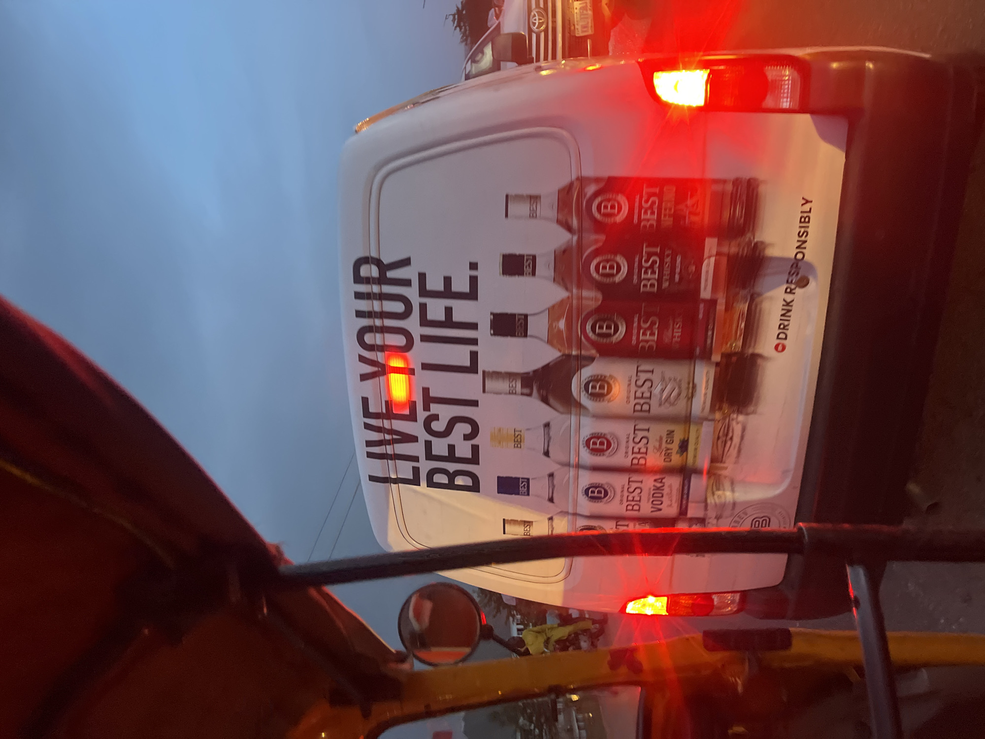 The image shows the rear of a white van with an advertisement for "Best" brand alcoholic beverages. The advertisement prominently features the text "LIVE YOUR BEST LIFE" and rows of bottles labeled "BEST" Vodka, Dry Gin, Whiskey, and Inferno Whiskey. The tagline "DRINK RESPONSIBLY" is visible in a smaller font. The van's brake lights are illuminated. The photo appears to have been taken from within a yellow vehicle, possibly an auto-rickshaw, based on the visible frame and side mirror. The location is unknown.