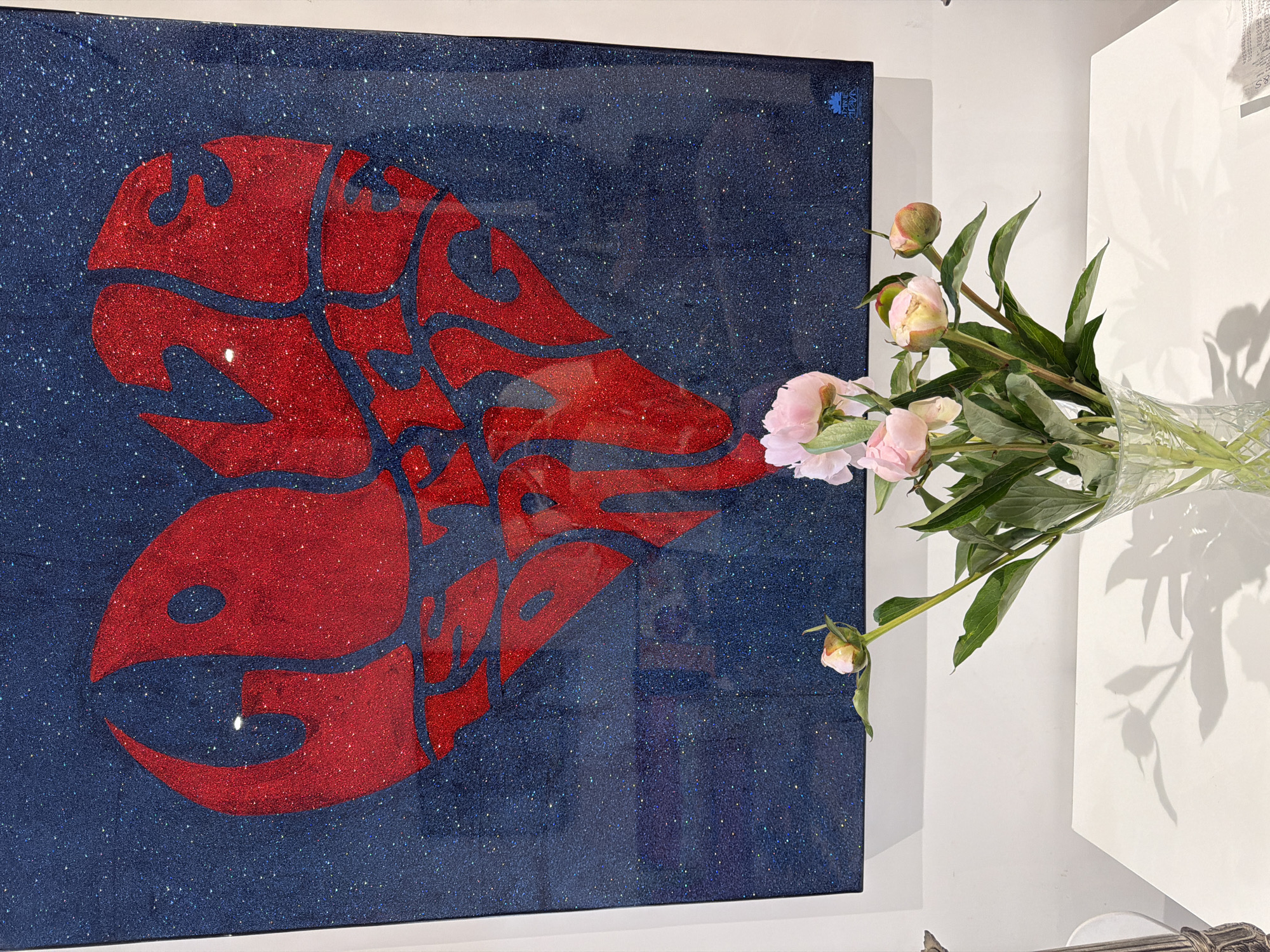 The image depicts a framed artwork, possibly a painting or a mixed media piece, displayed next to a vase of pink peonies. The artwork features a stylized, abstract design in red against a background of deep blue with glittery accents, reminiscent of a star-filled night sky. The design resembles a lobster or crustacean motif. The artwork is hung on a white wall in a location which, although not visible, is inferred to be in London, United Kingdom based on the instructions. A clear glass vase containing the pink peonies stands to the right of the artwork. The lighting suggests an interior space, with a strong shadow cast by the flowers on the white wall. No people are visible in the frame.