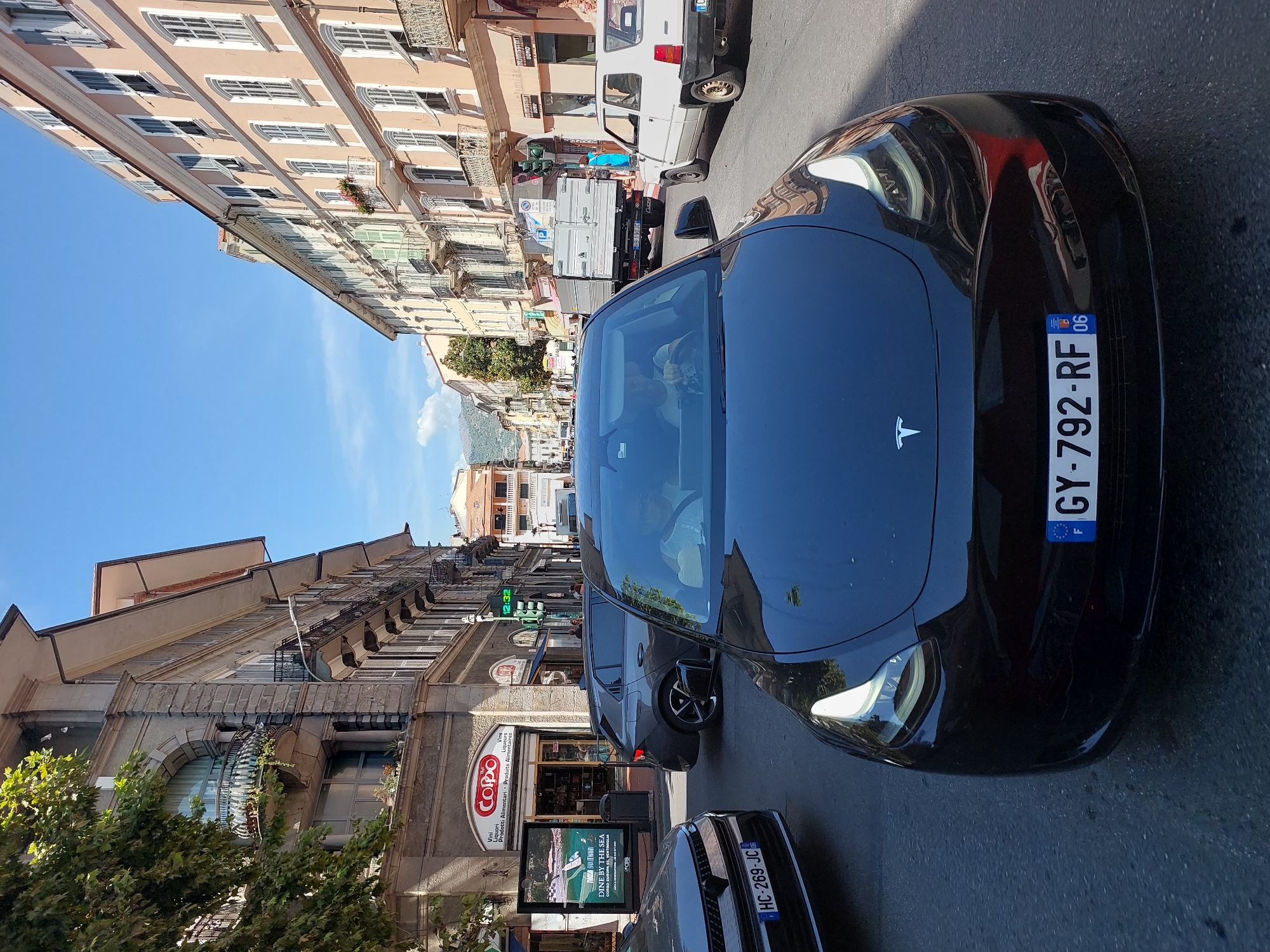 The image captures a street scene in Ventimiglia, Italy, on a bright, sunny day. In the foreground, a black Tesla Model 3 is prominently featured, facing the camera and driving down the street. The car's license plate reads "GY-792-RF 06". Inside the Tesla, two individuals are visible, a driver and a passenger, both appearing to be looking forward.
To the left of the Tesla, a dark-colored car with the license plate "HC-269-JC" is partially visible. Further to the left, a grey SUV is stopped. Behind the SUV, a building with a red sign reading "CORPO" is visible, indicating a shop. An advertising billboard is also present next to the shop.
The street is lined with multi-story buildings on both sides, characteristic of an Italian urban environment. These buildings feature balconies, arched windows, and traditional architecture. The street itself is paved with asphalt. In the mid-ground, a white Fiat Panda is parked on the right side of the street, and a flatbed truck is visible further down the road. The sky is clear blue with a few faint clouds. The overall atmosphere suggests a bustling daytime scene in a European city.