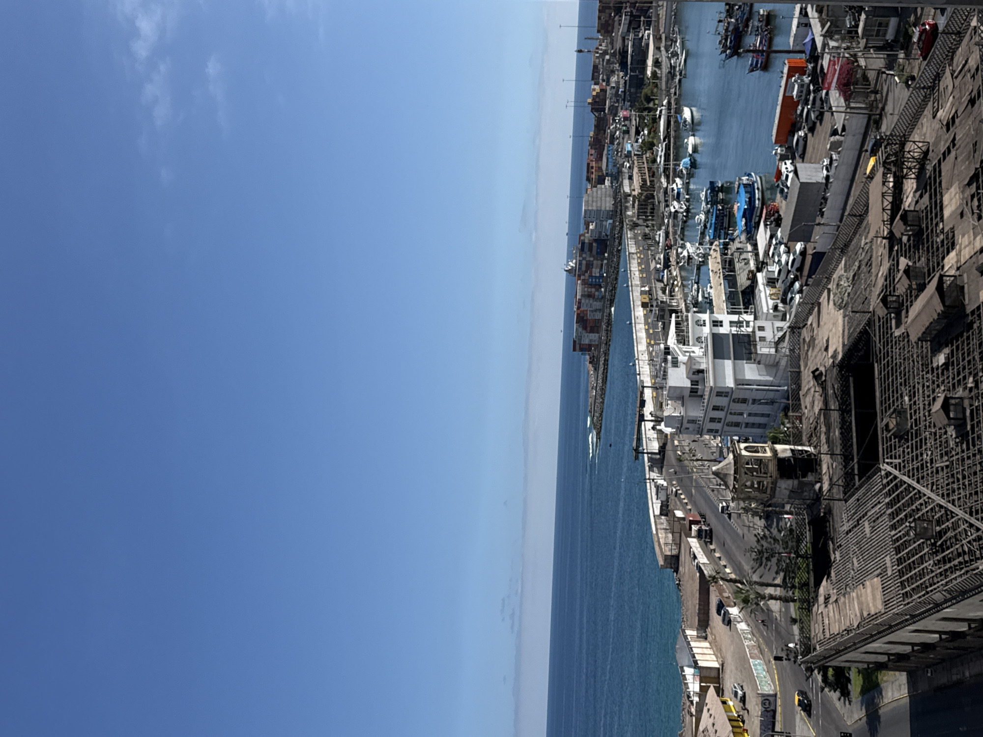 The image captures a view of Iquique, Chile, with the Pacific Ocean on the left and the cityscape on the right. The foreground features the damaged roof of a building, revealing its wooden structure. Adjacent to this, a road runs through the city, bordered by palm trees and a mix of older and modern buildings. In the midground, a white building with multiple stories stands prominently. Further into the scene, a harbor is visible, filled with various boats and yachts. Across the harbor, a large cargo terminal is apparent, with numerous shipping containers stacked high and a large vessel in the water. The sky is clear and blue with a few scattered clouds. There are several vehicles on the road and parked in the vicinity of the harbor. No people are clearly distinguishable in the image.