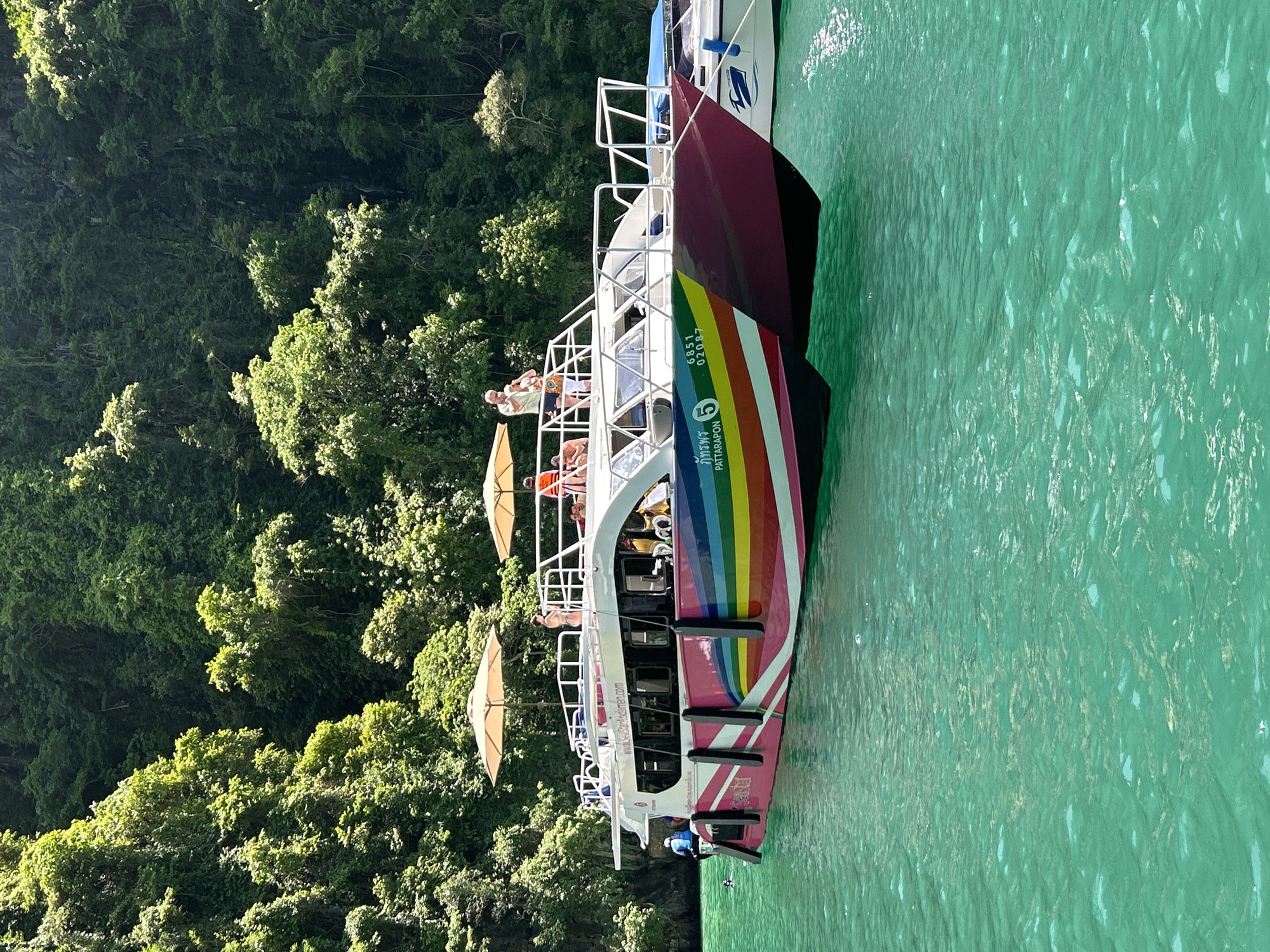 A pink and white catamaran, named "PATTARAPORN" (ภัทรพร) and numbered "5", is moored in clear, turquoise-green water within a cove in Ao Nang, Thailand. The catamaran features a distinctive rainbow stripe pattern across its hull, with additional identifying text "6851 02087" and the website "www.SeaStarAndaman.com" visible. On the upper deck, two large beige umbrellas are open, providing shade for passengers. Several individuals are present on the boat; on the upper deck, one person in a light shirt and shorts stands near an umbrella, while another in a bikini is nearby, and a third wears an orange life vest. A person in a blue life vest is partially submerged in the water at the stern of the boat, with another person's head visible in the water nearby. Black fenders line the side of the catamaran. To the right, the bow of a smaller white boat with blue markings is visible, also moored. The background consists of steep, verdant limestone karsts densely covered in lush green tropical vegetation, with sunlight illuminating portions of the foliage and casting others in shadow. The water exhibits minor ripples and reflections from the bright sun.