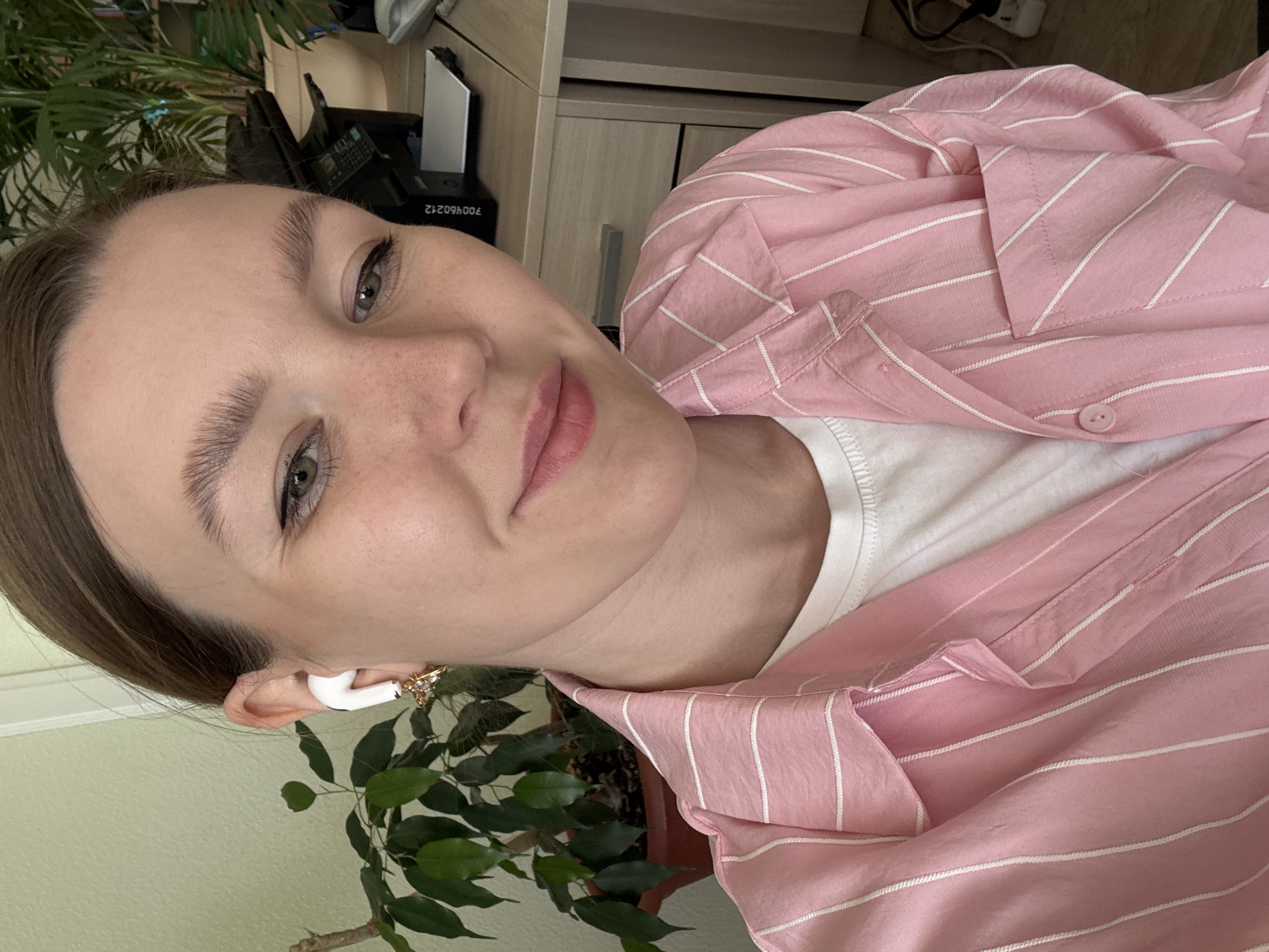 A person is depicted in a close-up, rotated selfie. The person has light skin, brown hair, and is wearing a white t-shirt and a pink and white striped button-down shirt. An earbud is visible in the left ear, and there is a piece of jewelry behind the ear. The background shows a green plant, a beige wall, and a desk with office equipment. The location appears to be an indoor office setting. There is no information available to identify the city as Izhevsk, Russia.