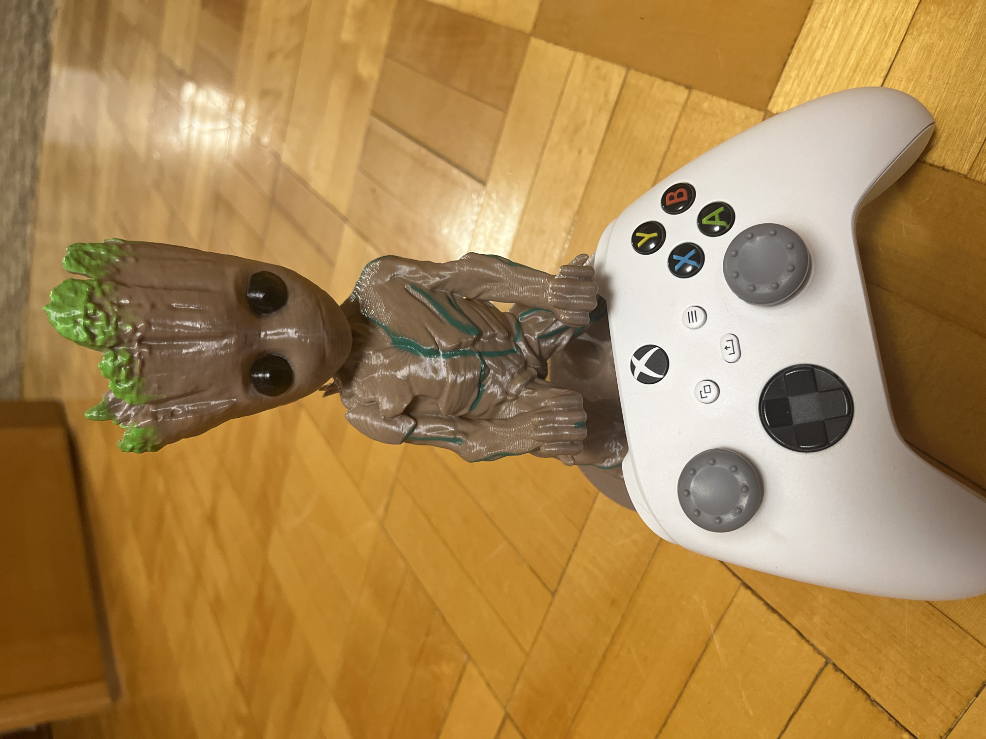 A 3D printed Groot figure is holding a white Xbox controller. The scene is set indoors on a wooden floor. The city and specific location are unknown.
