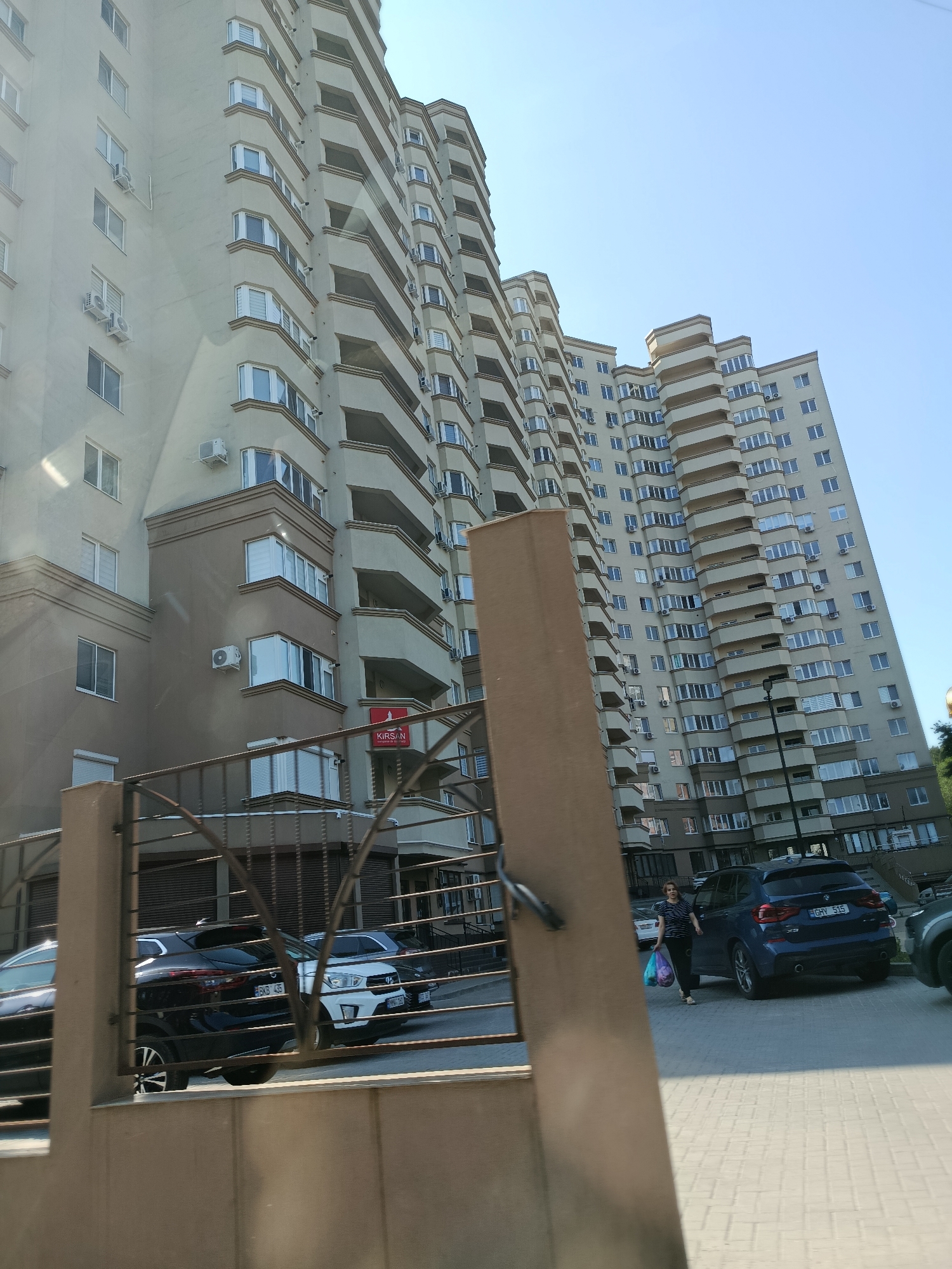 A woman is walking on a sidewalk in front of a large residential building in Chișinău, Moldova. The building is tall with many balconies and windows. There are cars parked in front of the building, and a fence in the foreground. The woman is carrying several shopping bags. The sky is clear and blue.