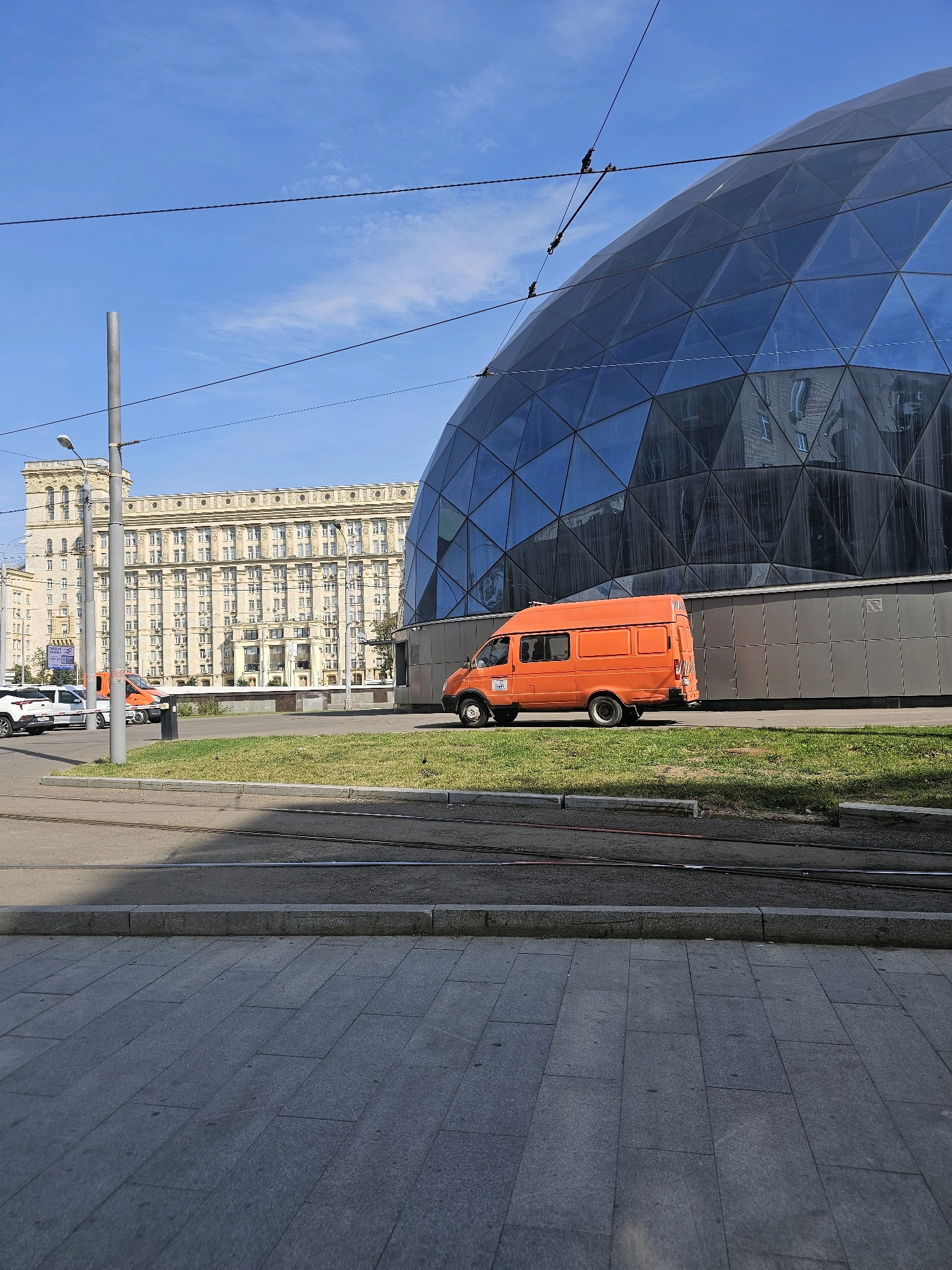 In Khimki, Russia, an orange van is parked on a grass median in front of a modern, geodesic dome-shaped building with a blue, faceted glass facade. To the left of the dome, a large, classical-style building with numerous windows stands under a clear blue sky. The foreground shows a paved walkway and tram tracks.