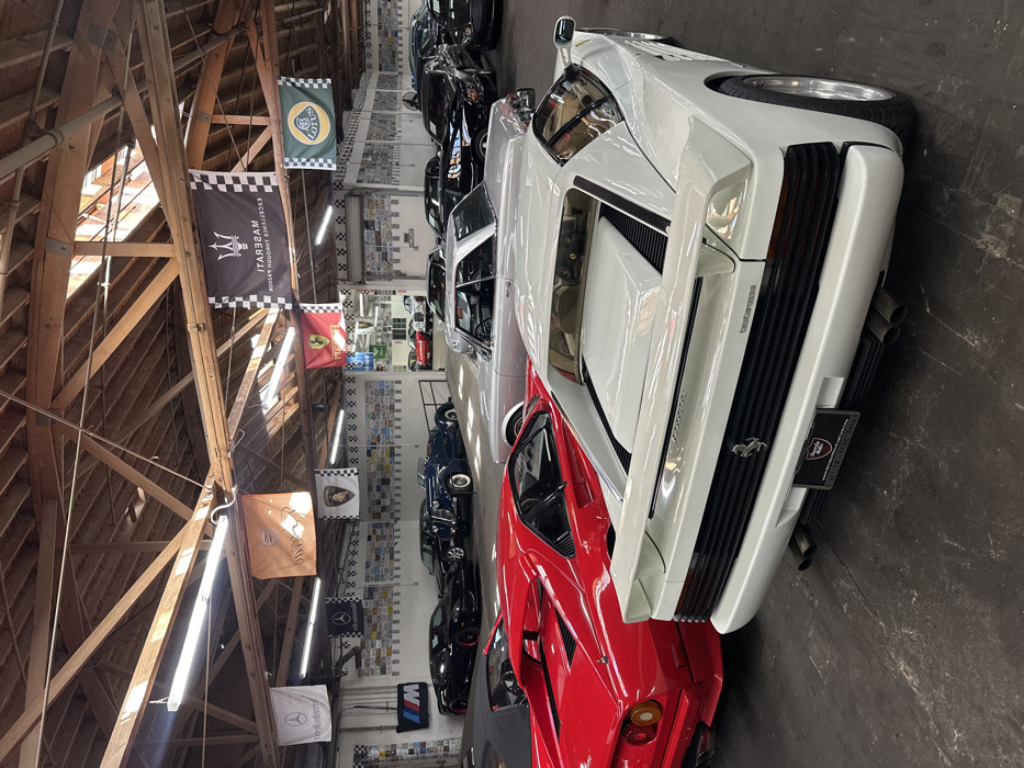 A collection of vintage and luxury cars lines a garage. A red Ferrari is nestled beside a white Testarossa with its signature black vents. Further down the line are other vehicles in shades of black, silver, and blue. Banners representing various automotive brands, including Lotus, Maserati, and Ferrari, hang from the wooden ceiling rafters. Numerous license plates adorn the wall, adding to the garage's automotive aesthetic.