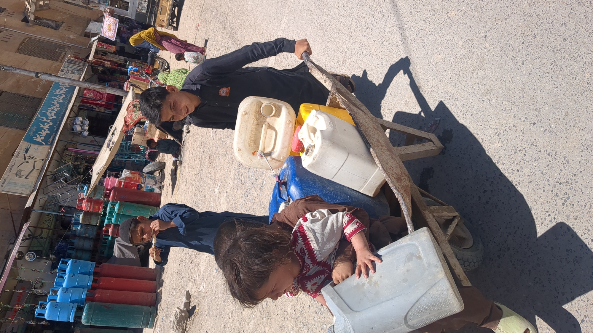 In Quetta City Tehsil, Pakistan, an adolescent boy in a long-sleeved black shirt pulls a wheelbarrow along a street. The wheelbarrow is filled with several plastic jerrycans of various colors and a young child wearing a brown outfit with red and white embroidery. Another boy, dressed in a dark blue shirt and traditional head covering, holds onto the side of the wheelbarrow. The scene is on a paved street, with shops and people visible in the background. Stacks of colored gas cylinders are displayed in front of one of the shops.
