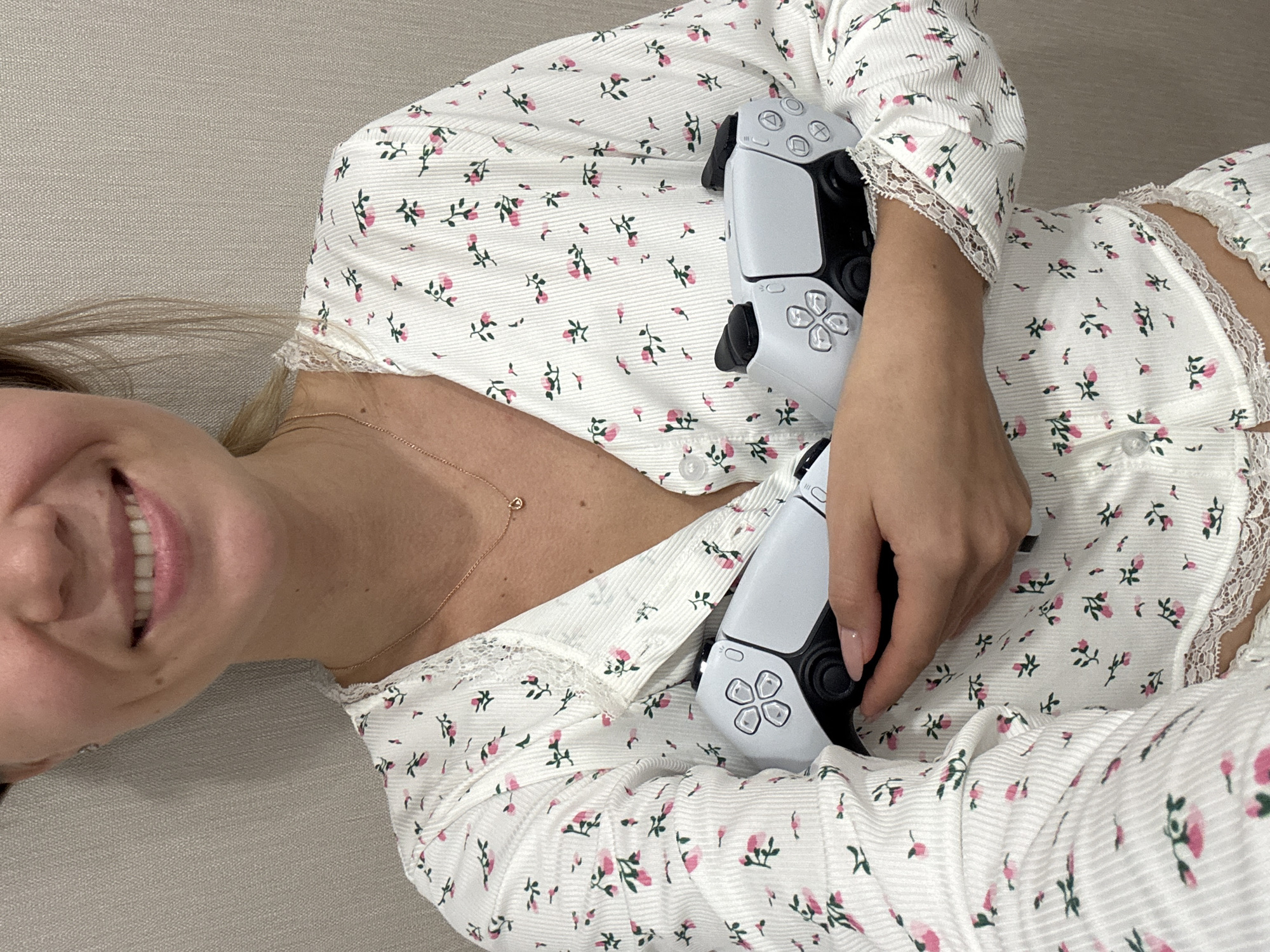 A woman in a floral pajama set is lying on what appears to be a light grey or beige surface. She is holding two white PlayStation 5 DualSense controllers. The controllers are positioned in front of her chest, with her hands visible holding them. She is smiling, and a gold necklace is visible around her neck. The environment is not discernible, and there is no city identified.