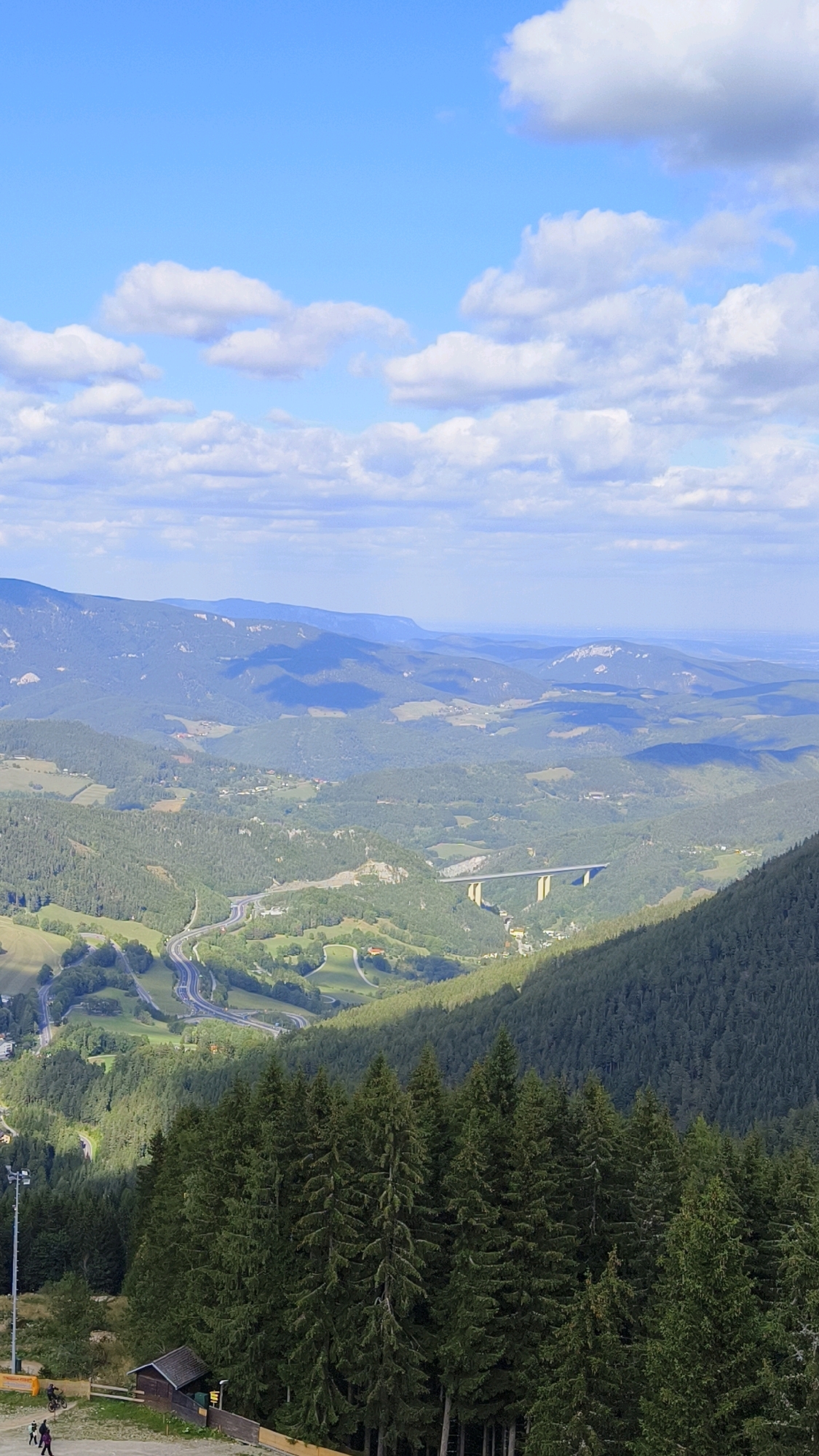 The image depicts a sweeping panoramic view of a mountainous landscape in Semmering, Austria. In the foreground, a dense forest of dark green pine trees dominates the lower portion of the frame, with a small, dark wooden structure visible at the bottom left, near a ski lift pole. A few people are walking or standing near the structure, and two figures on bicycles are also visible.
The mid-ground features a winding highway carved into the lush green hills, with several switchbacks visible. Further in the distance, a striking concrete bridge spans across a valley, with its piers supporting the roadway. The valley is dotted with scattered buildings and agricultural fields, creating a patchwork of greens and browns.
The background is characterized by rolling mountain ranges that recede into the distance, their slopes covered in dense forests. The mountains exhibit a hazy, bluish hue due to atmospheric perspective, with some lighter, rocky outcrops visible on the higher peaks. The sky above is a vibrant blue, punctuated by fluffy white clouds that cast shadows on the terrain below. The overall scene conveys a sense of natural beauty and the integration of human infrastructure within a rugged environment.