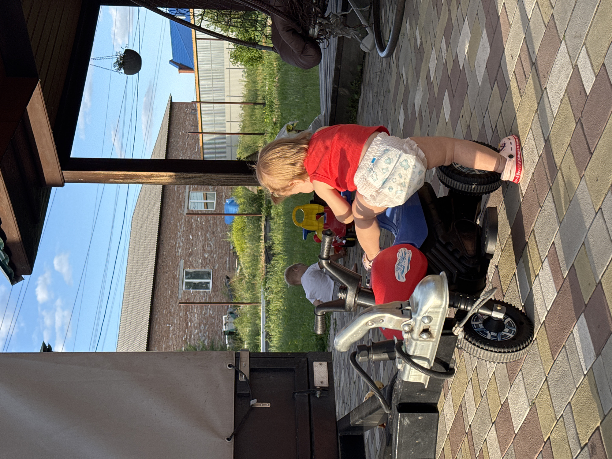 A young child wearing a red shirt and a diaper is playing with a toy motorcycle on a paved area. The child is leaning forward, with their hands on the handlebars. The motorcycle is red and silver, with a black wheel visible. In the background, there is a brick building, a grassy area, and a fence. A person is sitting in the grass to the left of the motorcycle. The scene appears to be outdoors, possibly in a yard or a playground, with dappled sunlight suggesting it is daytime. The sky is blue with some clouds. There is some text visible on the side of the child's shoe, but it is not clearly legible.