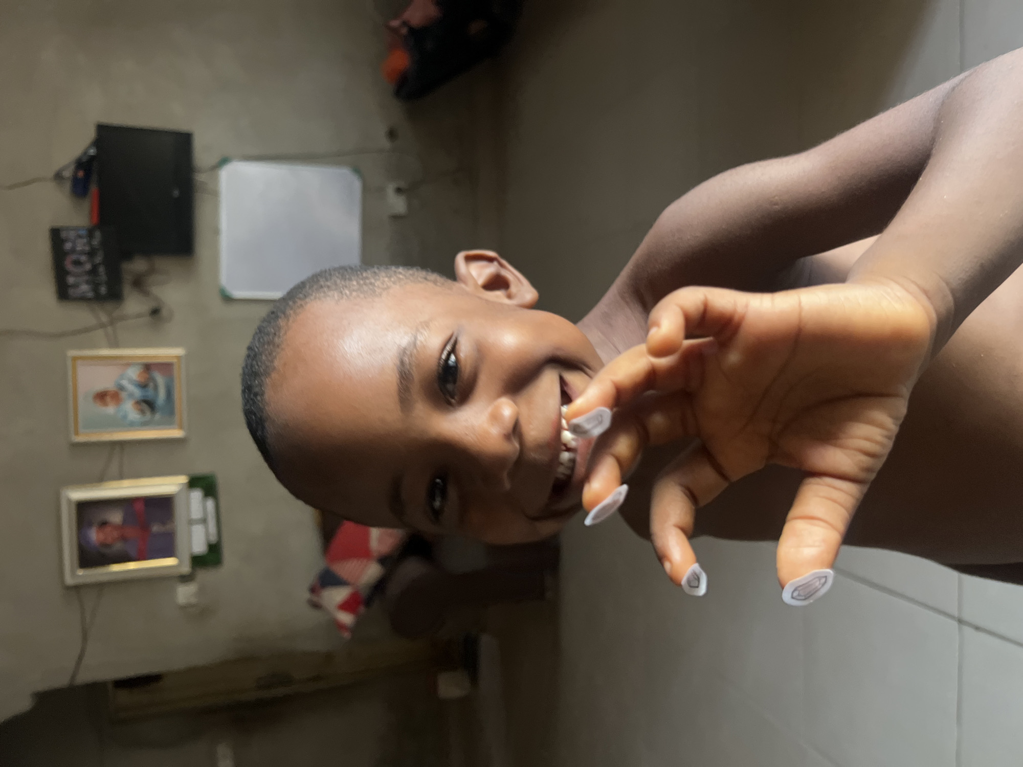 A young, dark-skinned child smiles at the camera, holding their hand up to show fingernails that are adorned with cartoon gem stickers. The background is a dimly lit indoor space, possibly a bedroom, with textured walls. Visible in the background are framed pictures hanging on the wall, a small whiteboard, and a black object that may be a monitor. The location is estimated to be in the city of Minna, Nigeria.