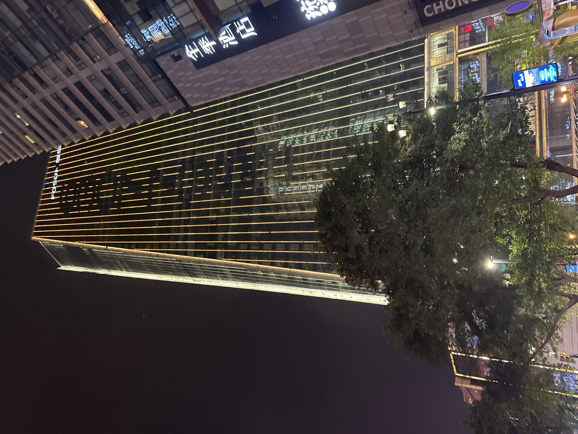 At night in Yuzhong District, Chongqing, China, a tall glass building with horizontal yellow lights is visible. The building is a modern structure with a sharp, triangular apex. Next to the building is a tree with dense green foliage. In the top right, another building is visible, with the word "CHONG" displayed on it. A street light illuminates the scene. There are no people visible in the image.