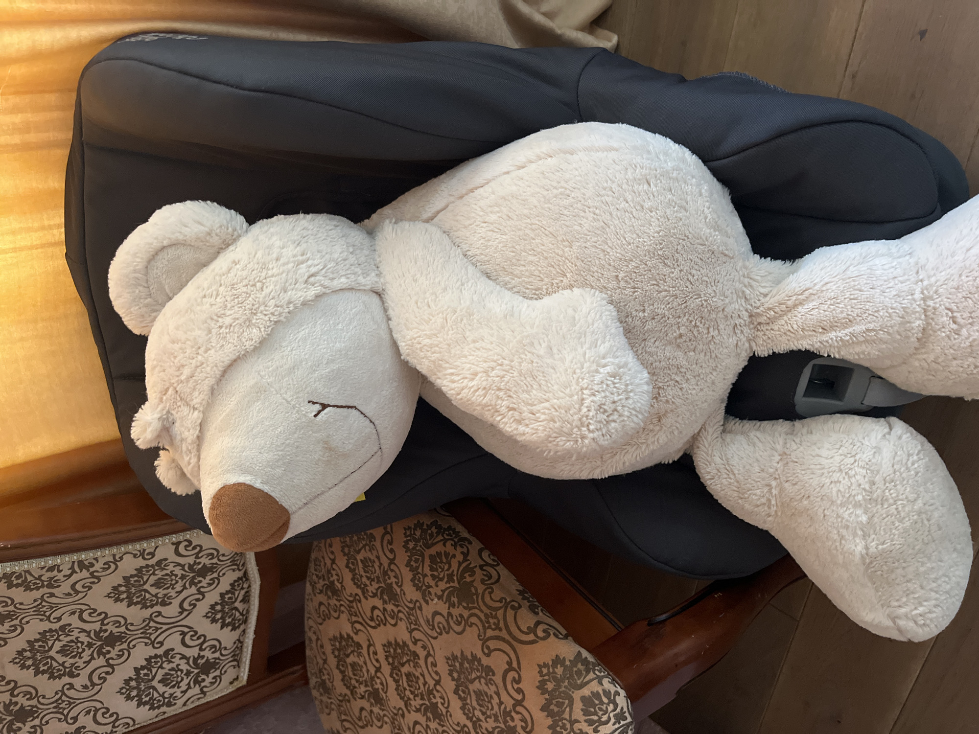 A light beige stuffed teddy bear is seated in a black car seat. The teddy bear is positioned facing left and is leaning against the car seat's headrest. Its right arm is bent and rests across its belly. The bear has a brown nose and stitched facial features. The car seat is in a room with wooden flooring and two chairs with patterned upholstery to the left. A yellow curtain is visible in the background. The scene is indoors, and there are no people present. The overall impression is one of domesticity. The image was taken in Tarnivtsi, Ukraine.