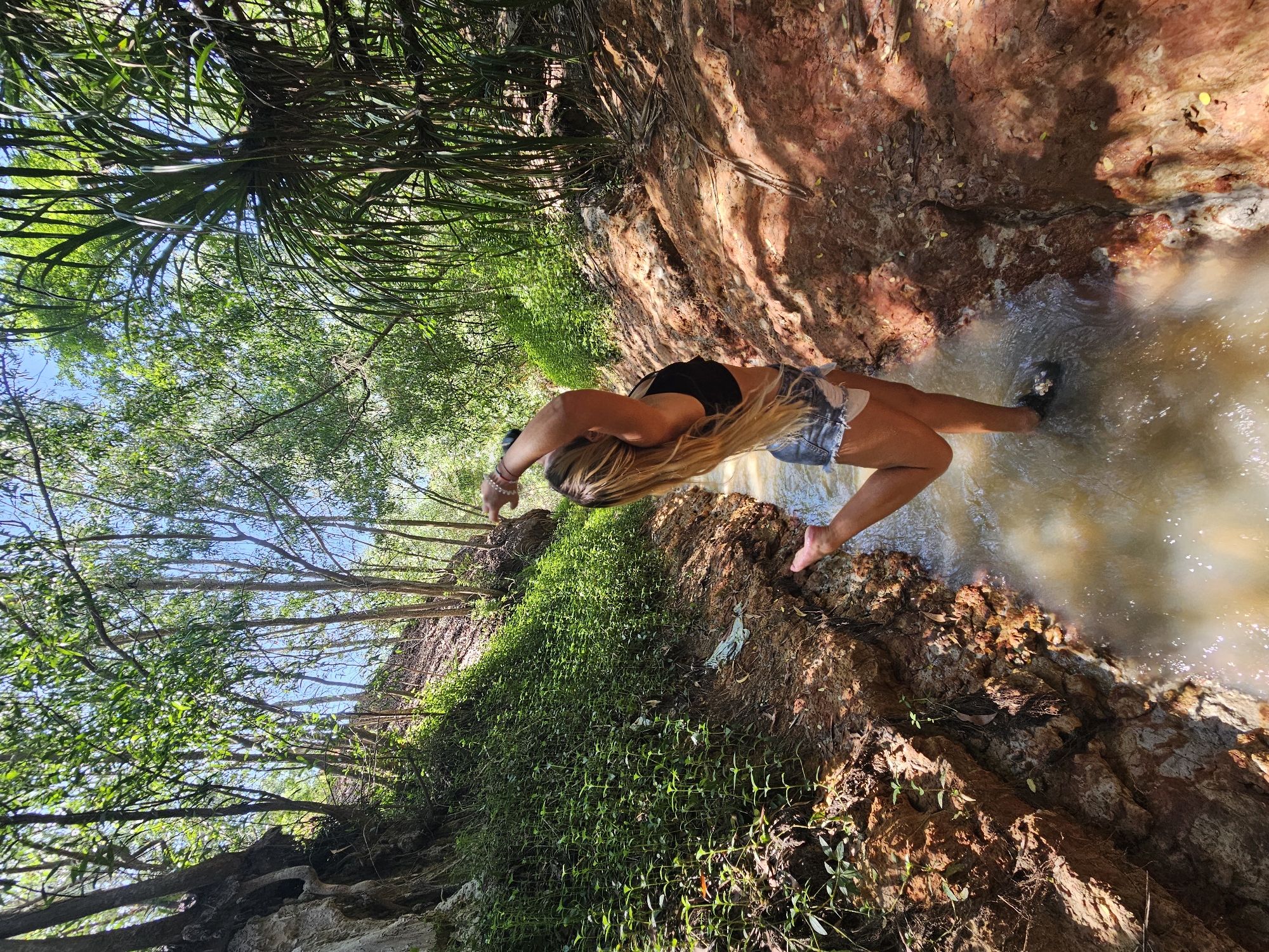 The media file shows a woman with long blonde hair, wearing a black top and denim shorts, standing in shallow, muddy water. She is positioned in the center of the frame, with her body arched backward and her arms extended. The setting appears to be a natural environment with a rocky, reddish-brown embankment on the right and lush green foliage and trees on the left. The water is murky and reflects the sunlight, creating shimmering patterns.
The scene suggests a moment of relaxation or enjoyment of nature, possibly a swim or wading in a stream or pool. The lighting indicates it is daytime, with bright sunlight casting shadows. The location, Mũi Né, Vietnam, is known for its beaches and sand dunes, but this scene suggests a more inland, perhaps a riverbed or a natural spring. The overall mood is serene and natural. There is no visible text in the image.