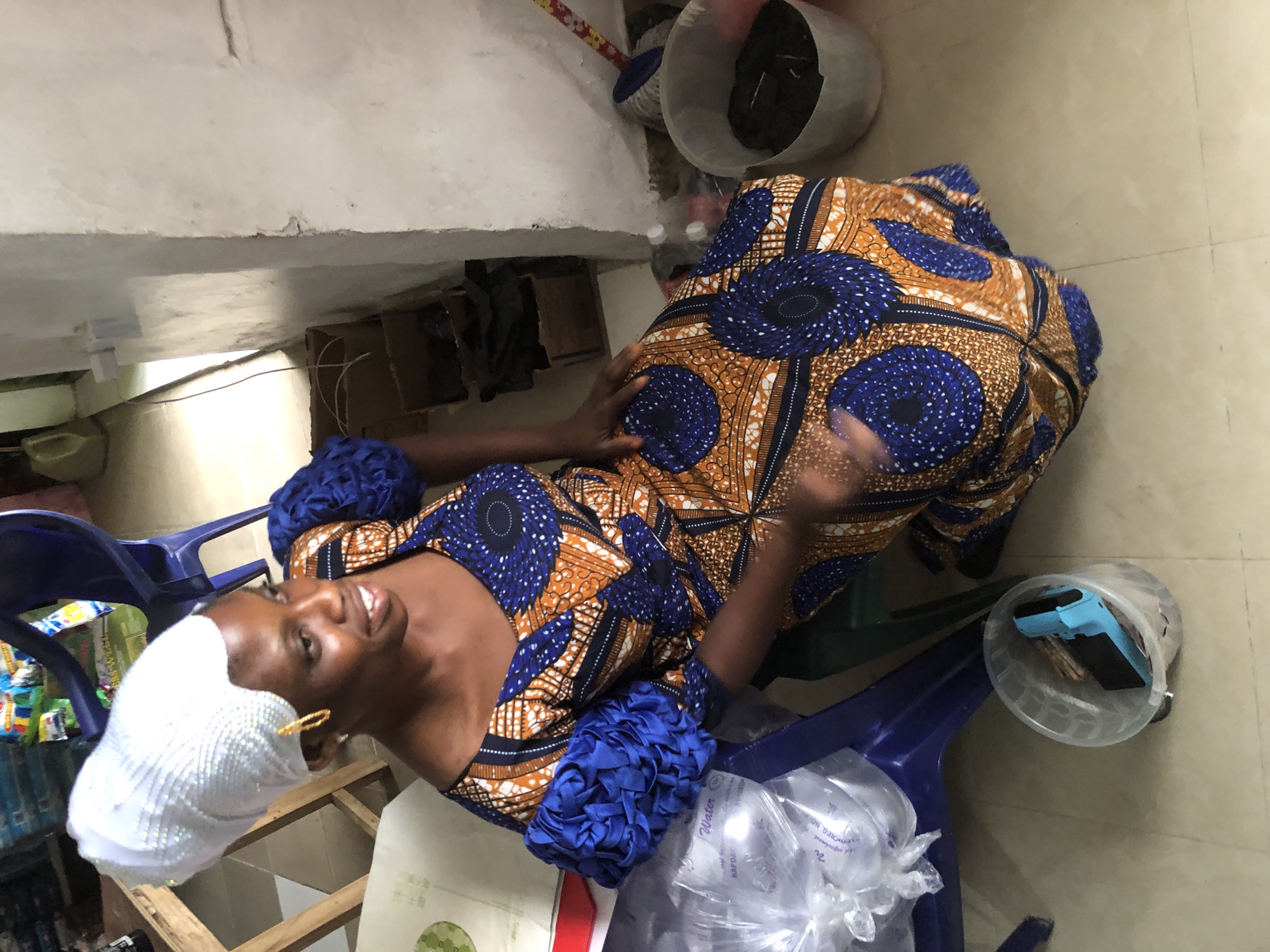 An overhead medium shot shows a woman sitting on a blue chair in what appears to be a room in Ado, Nigeria. The woman is wearing a white headwrap adorned with rhinestones and gold hoop earrings. She is dressed in a vibrant orange and blue patterned Ankara dress with puffed sleeves. Her skin tone is dark, and she is smiling as she looks to her left. Her right hand is resting on her lap, while her left hand is partially visible in motion.
The environment is cluttered. To the woman's left, a clear plastic bag filled with water bottles is on the floor. A clear plastic bucket containing dark material and a blue object is in the foreground to her right. Behind her, there are stacks of cardboard boxes and a blue plastic chair. In the background, a portion of a wooden table with items on it is visible, and a broom with a yellow and red handle leans against a white wall. The floor is tiled in a light beige color, and the wall to the right is a rough, textured white. The lighting suggests an indoor setting, possibly a workshop or storeroom.