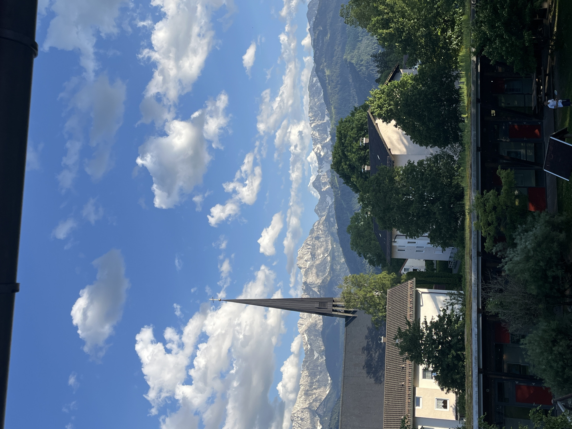 The image depicts a church spire and the Bavarian Alps against a cloudy blue sky in Garmisch-Partenkirchen, Germany. Buildings and trees fill the foreground on the right. In the top right, a seated person is visible in the far background.