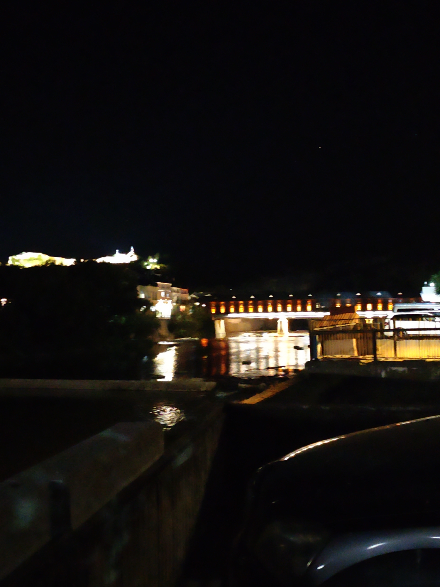 This is a night-time photograph taken from a vantage point overlooking the Osam River in Lovech, Bulgaria. The scene is dominated by the darkness of the night sky, with artificial lights illuminating sections of the cityscape.
In the background, a prominent hill features a cluster of illuminated buildings, appearing as white and yellow lights against the dark silhouette of the terrain. This likely represents the historic Varosha district or the covered bridge area.
A long, illuminated structure, possibly a bridge or a riverside promenade, stretches across the middle ground. It is lined with numerous glowing yellow lights, which are reflected in the water below. The river itself is dark, with shimmering reflections of the lights from the bridge and the opposite bank.
To the right of the bridge, a section of what appears to be a building or an outdoor area is brightly lit with yellow lights. A white vehicle, possibly a van or truck, is parked near this illuminated structure. A wooden fence-like barrier is also visible in this area.
In the foreground, a dark-colored car is partially visible on the right side of the frame, with its polished surface reflecting some of the ambient light. A concrete barrier or railing runs along the left side of the foreground, its surface textured and partially illuminated. The overall impression is one of a moderately lit urban environment at night, with the river serving as a central element.