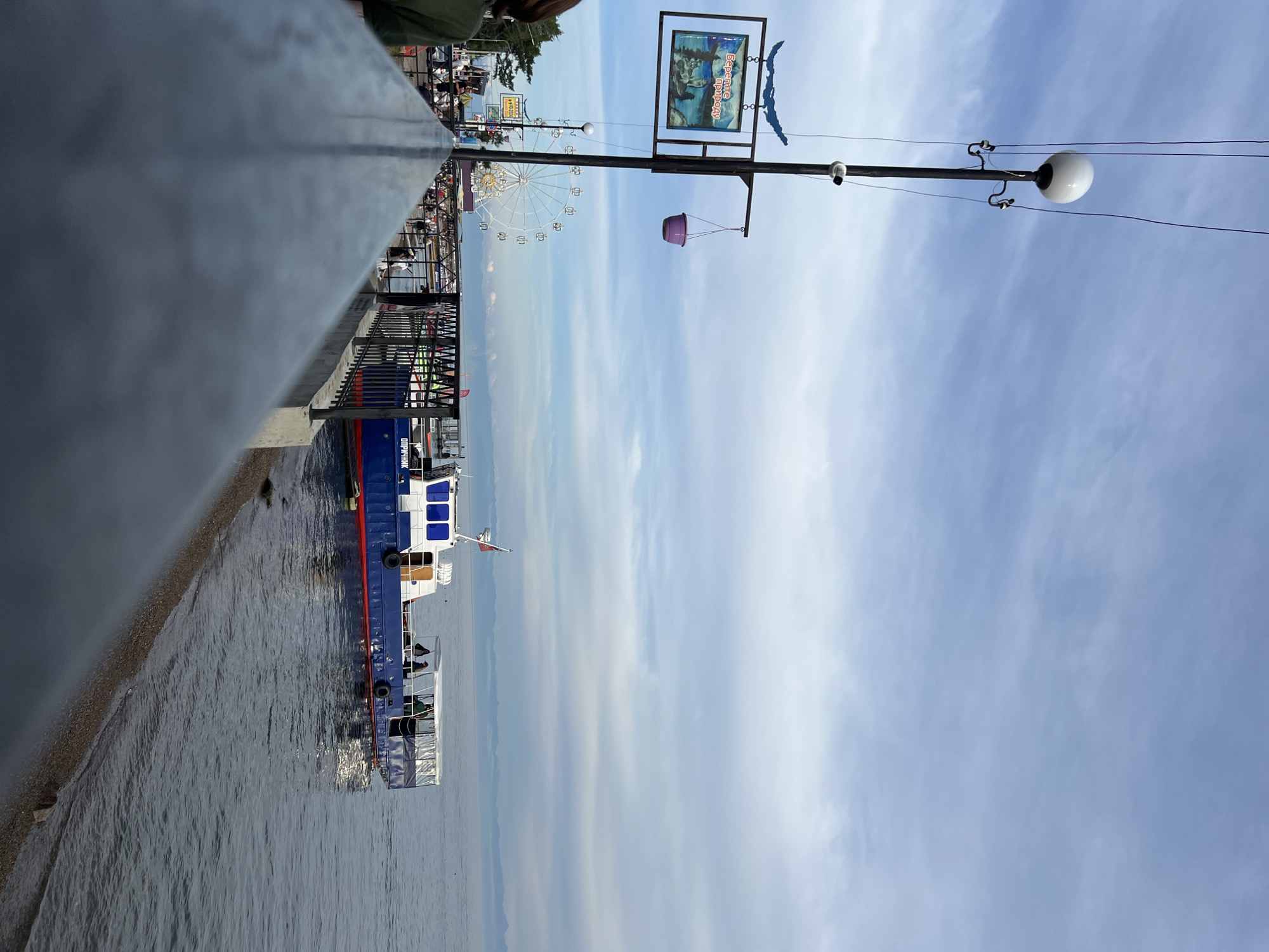 A blue and white boat is docked on a body of water. A pier runs along the foreground, with a metal railing. A Ferris wheel is visible in the background, along with a street lamp. A sign with Russian text is attached to the lamp post. The sky is cloudy. The scene appears to be in Listvyanka, Russia, a settlement on the southwestern shore of Lake Baikal.