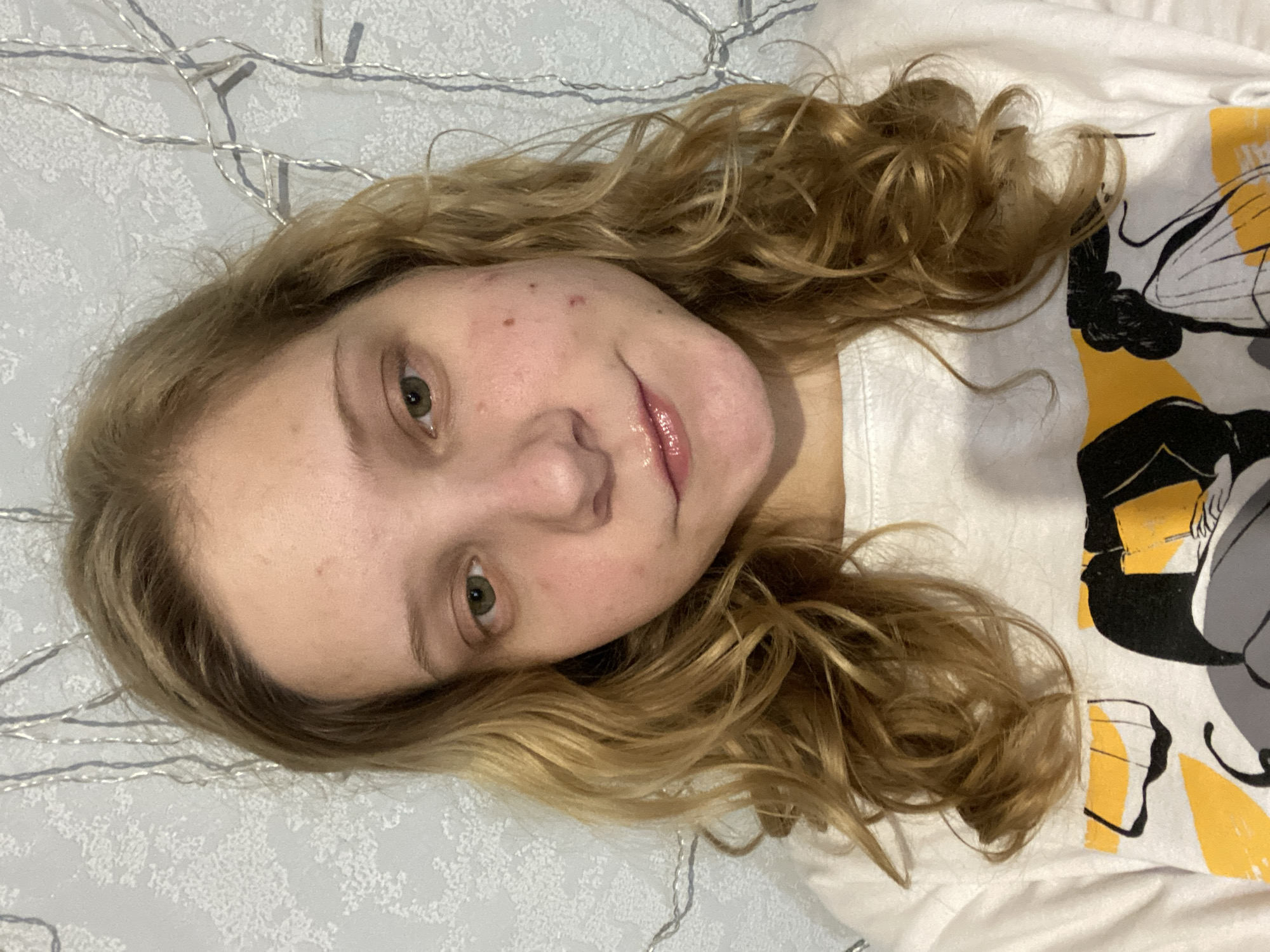 A young woman with fair skin and wavy, light brown hair is lying on a white textured surface. Her face is angled toward the camera, revealing green eyes and some mild acne. She is wearing a white t-shirt with a design featuring black and yellow elements. In the background, there are silver string lights. There is no evidence to determine the location as Chaikovsky, Russia.