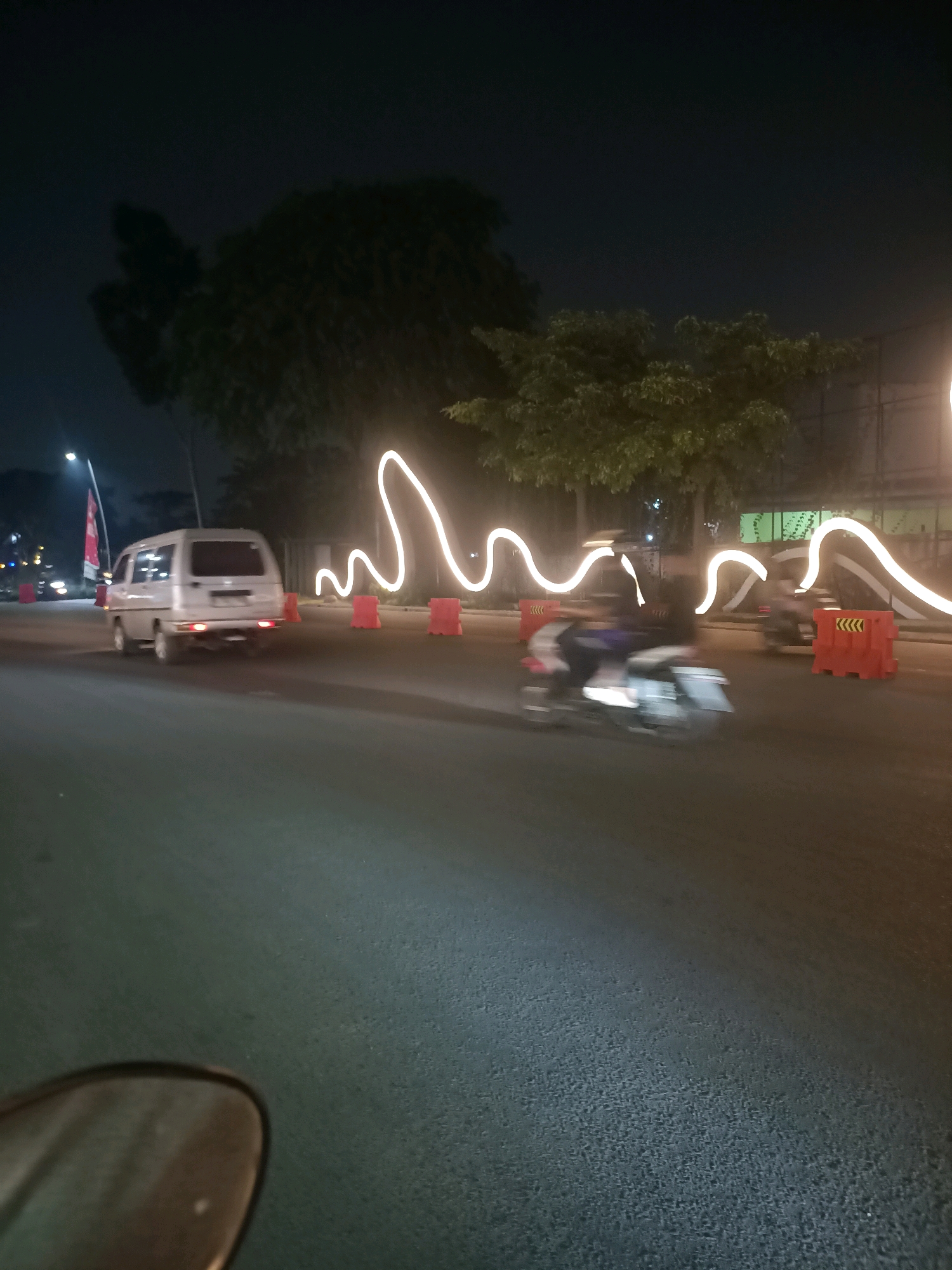 The image depicts a night scene on a road in Telagaasih, Indonesia. A silver van is moving away from the camera on the left side of the road. In the middle ground, a motorcycle is moving away from the camera, blurred by motion. Orange traffic cones with yellow chevron markings are visible along the side of the road. A wavy line of bright white lights, possibly a decorative light installation, runs along the median. Trees and streetlights are present in the background. The road surface is asphalt. The perspective suggests the photo was taken from another moving vehicle.