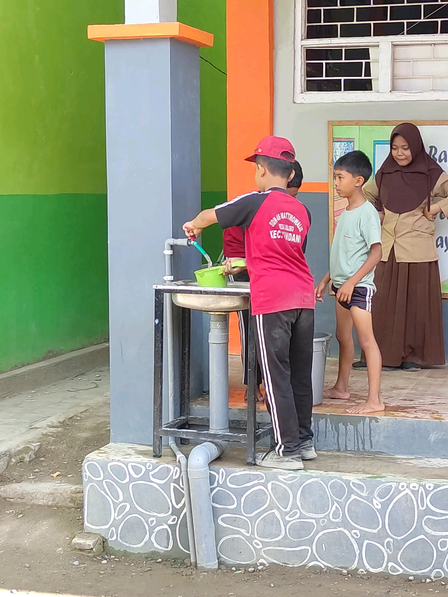 In Bulukumba, Indonesia, two young boys stand near a sink outside. One boy, wearing a red shirt and cap, turns on a faucet, filling a green bucket. The other boy, in a light blue t-shirt and dark shorts, stands barefoot and watches. Behind them, a woman in a brown hijab and school uniform stands with a poster. The scene appears to be near a school, with a green wall and a pillar visible.