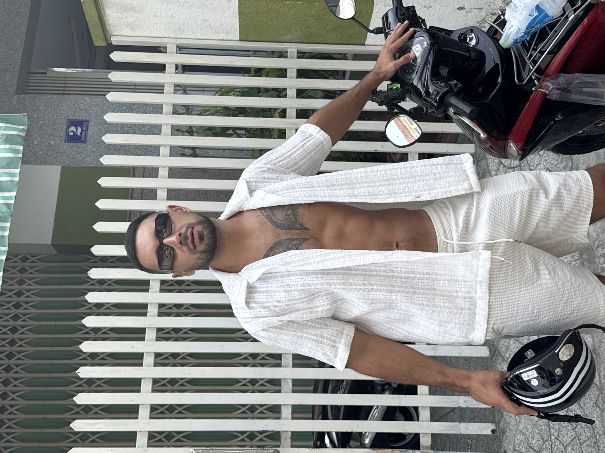 A man stands next to a red and black motorcycle on a city street in Nha Trang Ward, Vietnam. He is wearing a white short-sleeved open collar shirt, revealing his bare chest with a tattoo. He also wears white shorts and black sunglasses. He holds a black motorcycle helmet in his left hand. The background features a white picket fence and a building with a green stripe. A blue street sign with the number 2 is visible on the building.