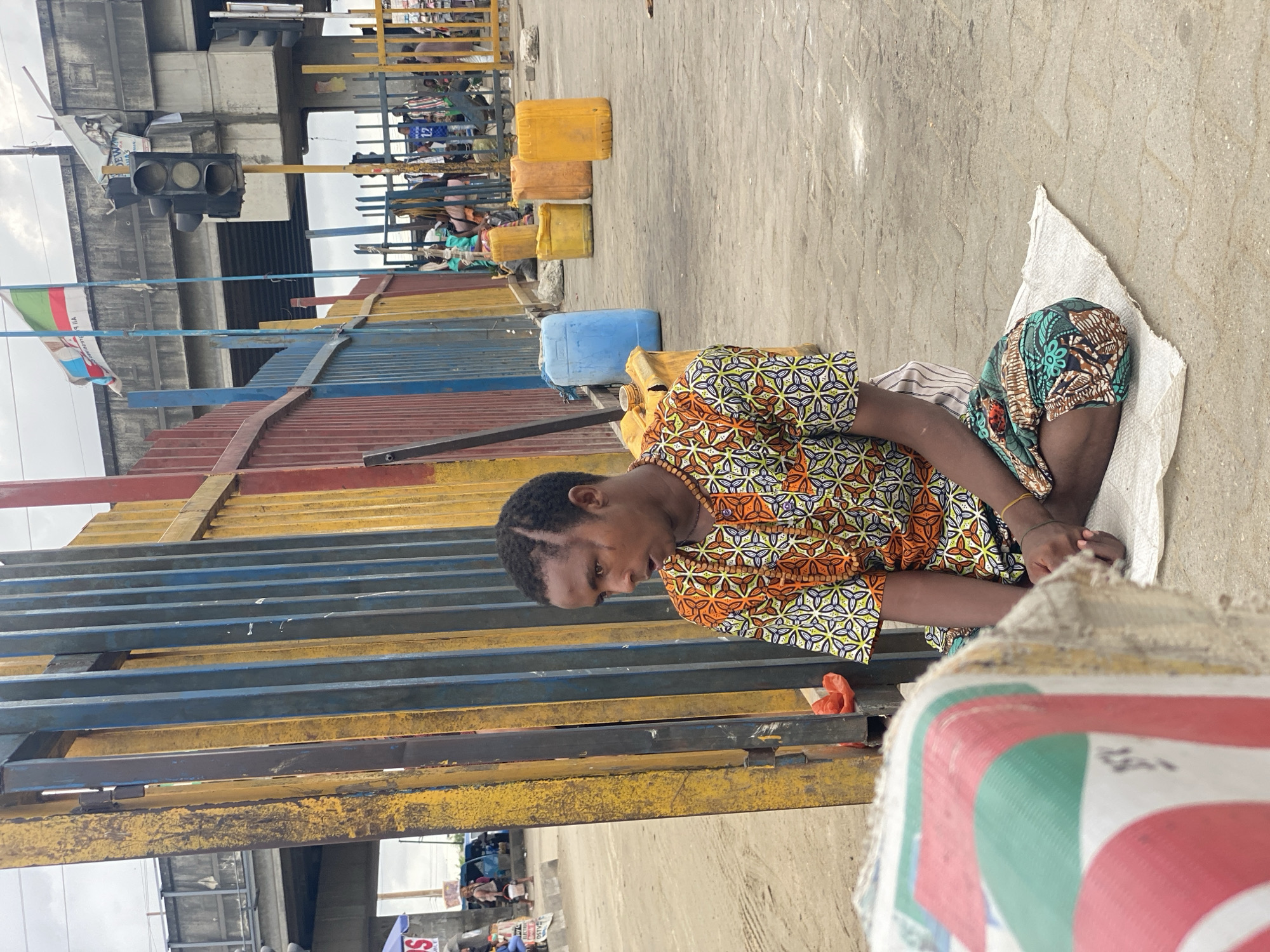 The image captures an outdoor scene in what appears to be a busy urban setting in Lagos, Nigeria. The central subject is a woman sitting on a white woven mat on a paved concrete ground, positioned slightly to the right of the frame. She wears a vibrant, short-sleeved shirt with an intricate geometric pattern in orange, green, yellow, and blue hues, and a dark green patterned skirt or wrapper. A wooden bead necklace adorns her neck. Her expression is calm, as she looks downwards.
Behind the woman, several yellow and one blue plastic jerrycans are neatly lined up on the ground. A multi-colored barrier or fence, constructed from horizontal metal bars in yellow, blue, and reddish-brown, extends across the middle ground, creating a visual separation. Beyond this barrier, blurred figures of other people suggest a crowded area, possibly a market or transportation hub.
In the upper left portion of the image, a black traffic light is mounted on a pole, and a multi-colored flag with horizontal stripes, including green, white, and red, is flying. Large, gray concrete structures are also visible in the background, likely part of an overpass or building. The sky appears overcast, indicating daytime. The scene portrays a typical street environment, characterized by functional objects, active human presence, and everyday urban elements. No distinct readable text is clearly visible on signs or objects, though some faint patterns are on the white mat she sits on.