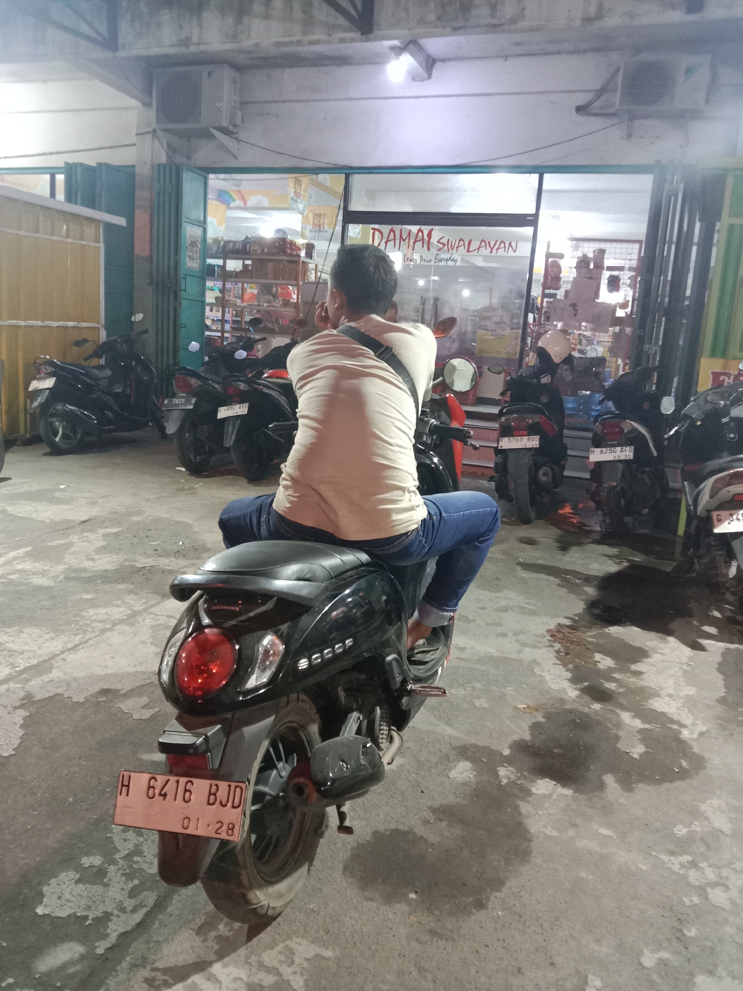 A man sits on a black scooter facing away from the camera in front of a store. He is wearing a tan shirt and blue jeans. The scooter has a license plate that reads "H 6416 BJD 01.28". Behind the scooter, several other motorcycles are parked, and a store with the name "DAMAI SWALAYAN" is visible. The store appears to be a convenience store or small supermarket. The ground is concrete and wet in some areas, possibly from rain or spillage. The lighting suggests it is evening or nighttime. The image is taken from a medium distance.