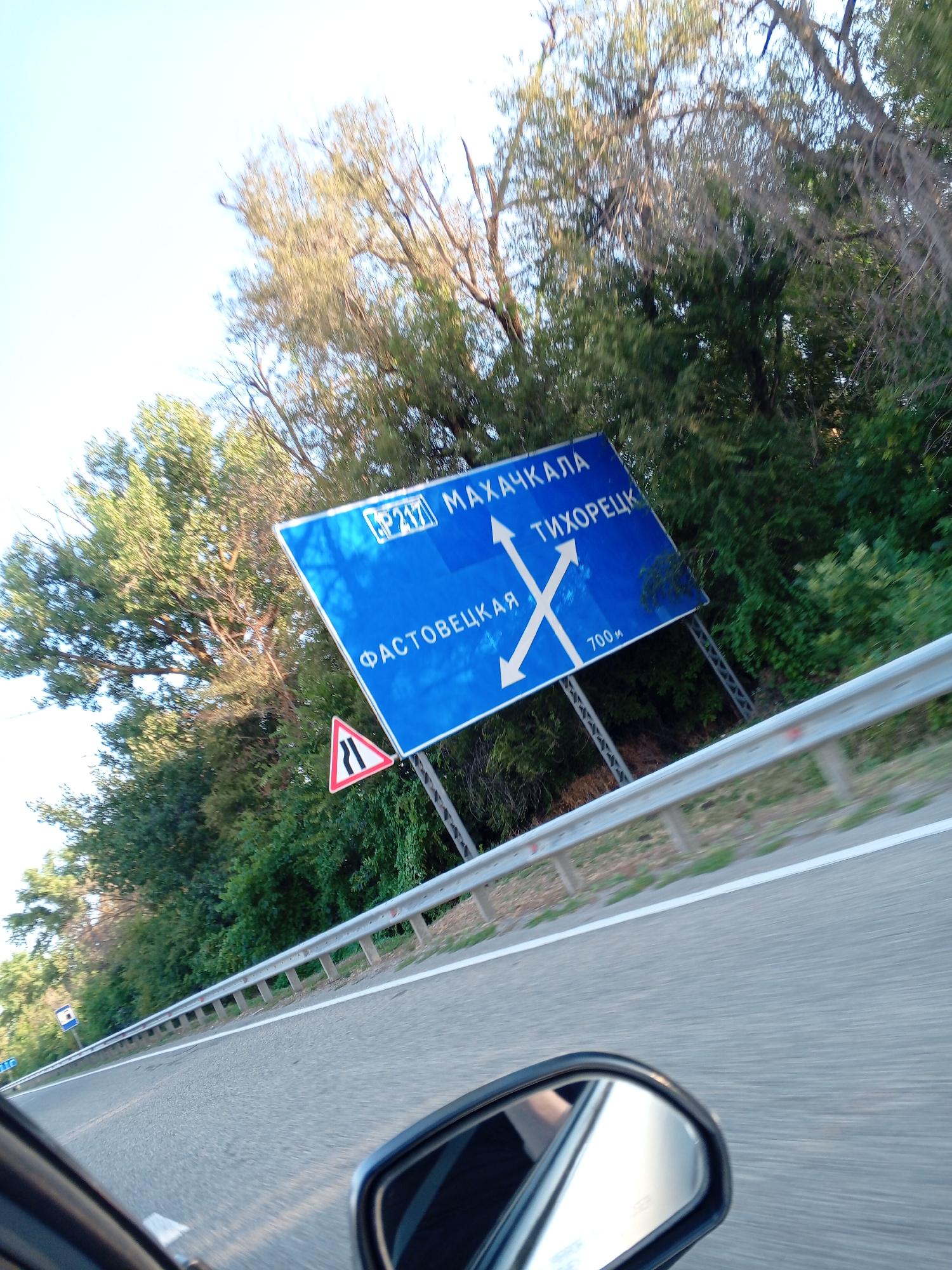 The image captures a tilted perspective of a blue directional road sign against a backdrop of lush green trees and a bright sky. The sign is triangular and displays white text and arrows, indicating directions to "МАХАЧКАЛА", "ТИХОРЕЦК", and "ФАСТОВЕЦКАЯ". There are also route numbers "P217" and a distance marker "700м" on the sign. Below the main sign, a smaller triangular red and white warning sign with a black symbol is visible. The road is visible in the foreground, with a metal guardrail running along its side. A reflection of the scene, including the driver's side mirror, is present in the lower right corner of the image. The overall impression is of a road trip or travel scene.