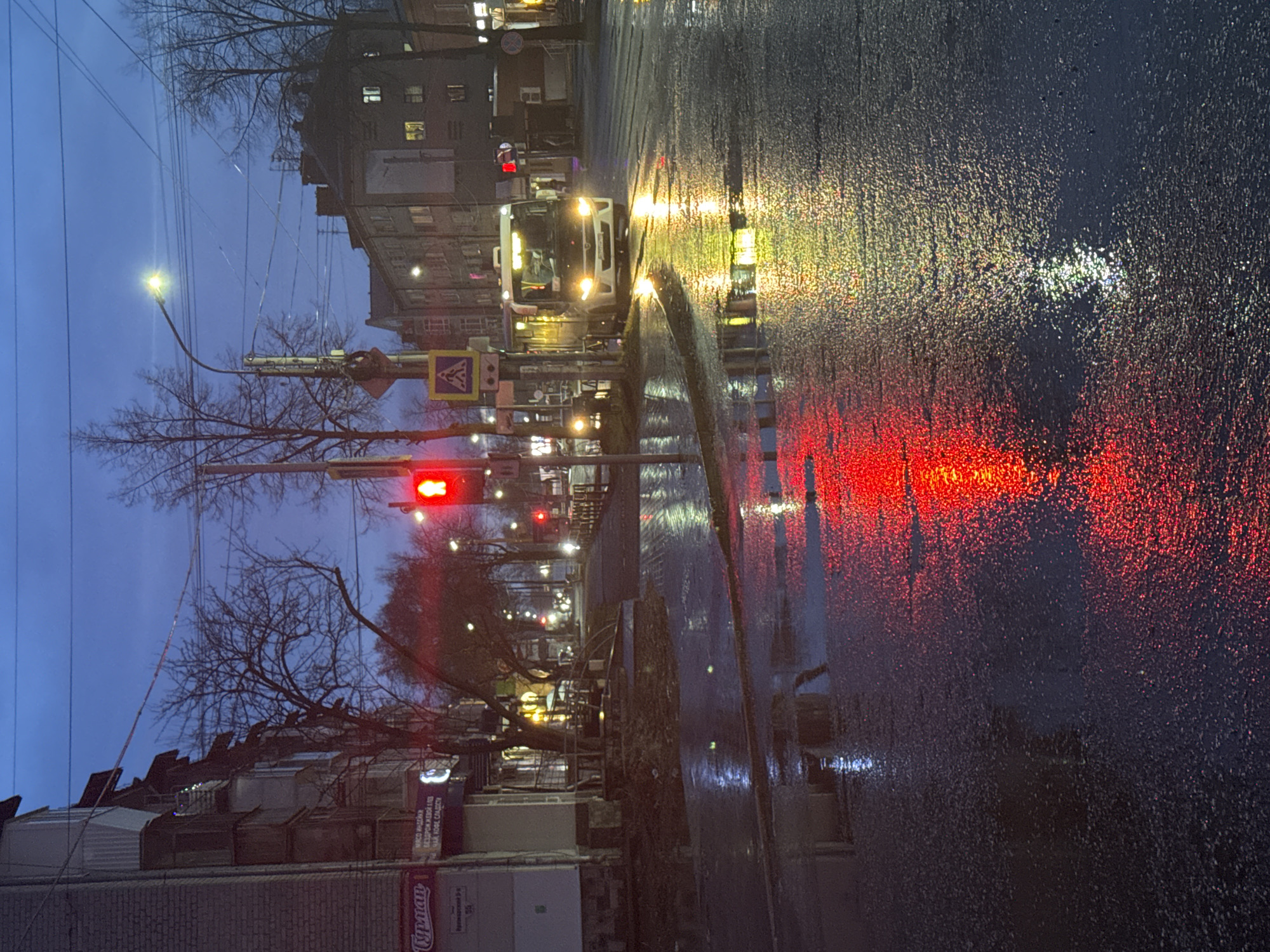 The media file captures an evening scene on a wet city street in Chaikovsky, Russia. The sky is a deep, dark blue, indicating dusk or nightfall. The street is visibly wet from recent rain, creating numerous vibrant reflections of artificial lights.
In the mid-ground, a white bus is prominently featured, stopped at a red traffic light. Its headlights are brightly illuminated, casting a strong, yellowish reflection on the wet road surface. The red traffic light itself is visible on the left side of the street, and its vivid red light creates a dominant, elongated reflection across the foreground of the wet asphalt. Other streetlights and lights from buildings also reflect, appearing as bright streaks and pools of light.
Multi-story buildings line both sides of the street, some with illuminated windows. Bare trees with intricate branch patterns are visible along the sidewalks, silhouetted against the sky and various light sources. A yellow triangular sign with a black pedestrian symbol, indicating a pedestrian crossing, is mounted on a pole near the traffic light. Utility poles and a network of power lines are visible overhead.
Visible text includes Cyrillic script on a sign below the pedestrian crossing, likely indicating a bus stop. On a billboard or sign in the lower-left, the Cyrillic word "СИМПЛЕМЕН" is discernible, along with partial text that includes "ЧАЙ". The scene evokes the atmosphere of a busy urban environment after rain, with the wet surfaces enhancing the visual impact of the city lights.
