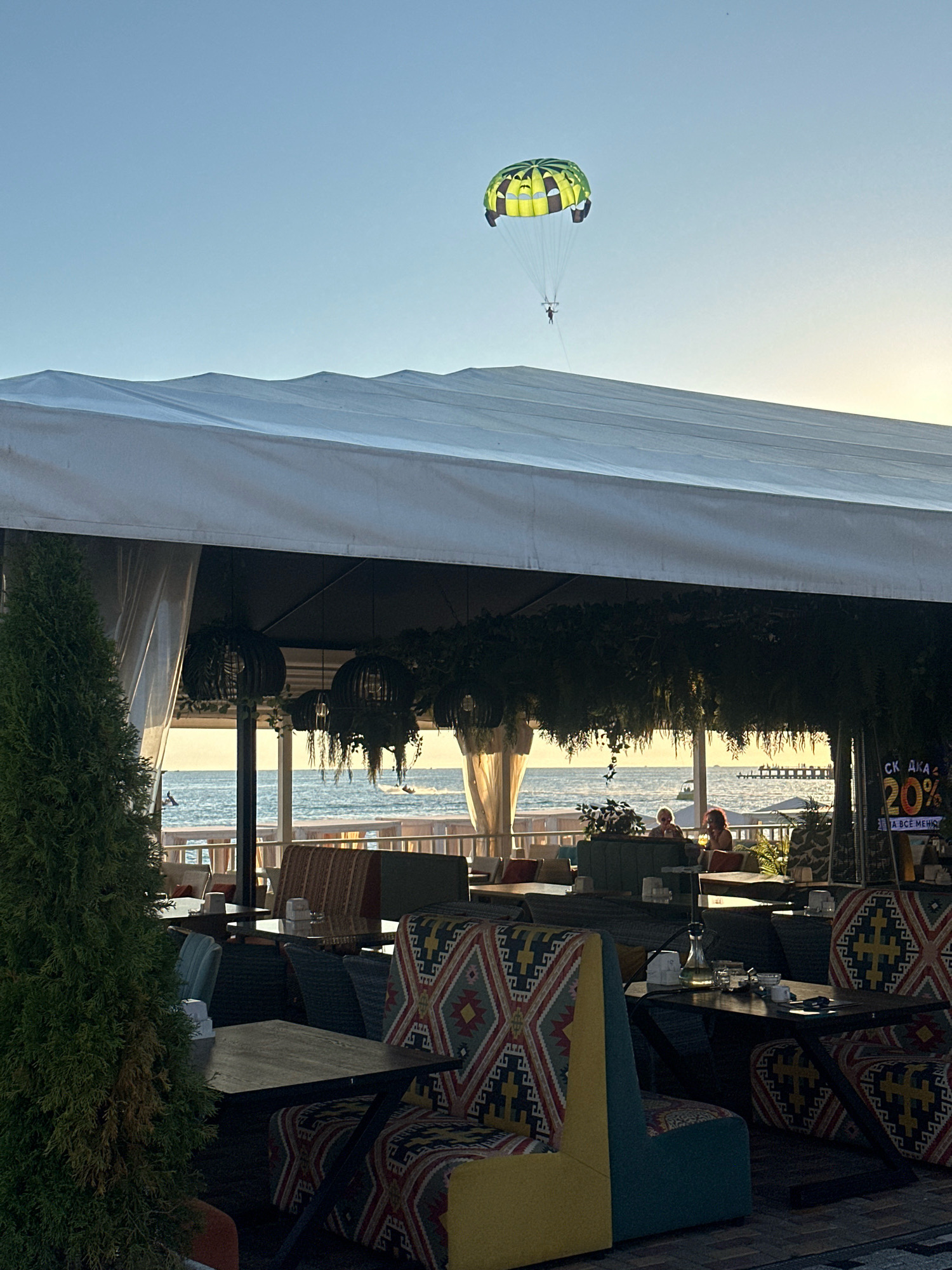 The image depicts an outdoor cafe or restaurant area overlooking the Black Sea in Lermontovo, Russia. In the foreground, there are tables and seating arrangements, some with vibrantly patterned upholstery. A large white canopy covers the seating area. To the left, a tall evergreen tree is visible.
In the middle ground, beyond the seating, a wooden deck or boardwalk separates the cafe from the sea. Several patrons are seated at tables, enjoying the view. The Black Sea stretches out to the horizon, with a pier visible in the distance.
In the sky above, a person is parasailing, attached to a bright green and yellow parachute. The sky is clear with a gentle gradient of blue. The overall atmosphere suggests a relaxed, seaside dining experience, likely during the late afternoon or early evening given the lighting. A sign with Cyrillic text advertising a "20%" discount is partially visible on the right side of the image.