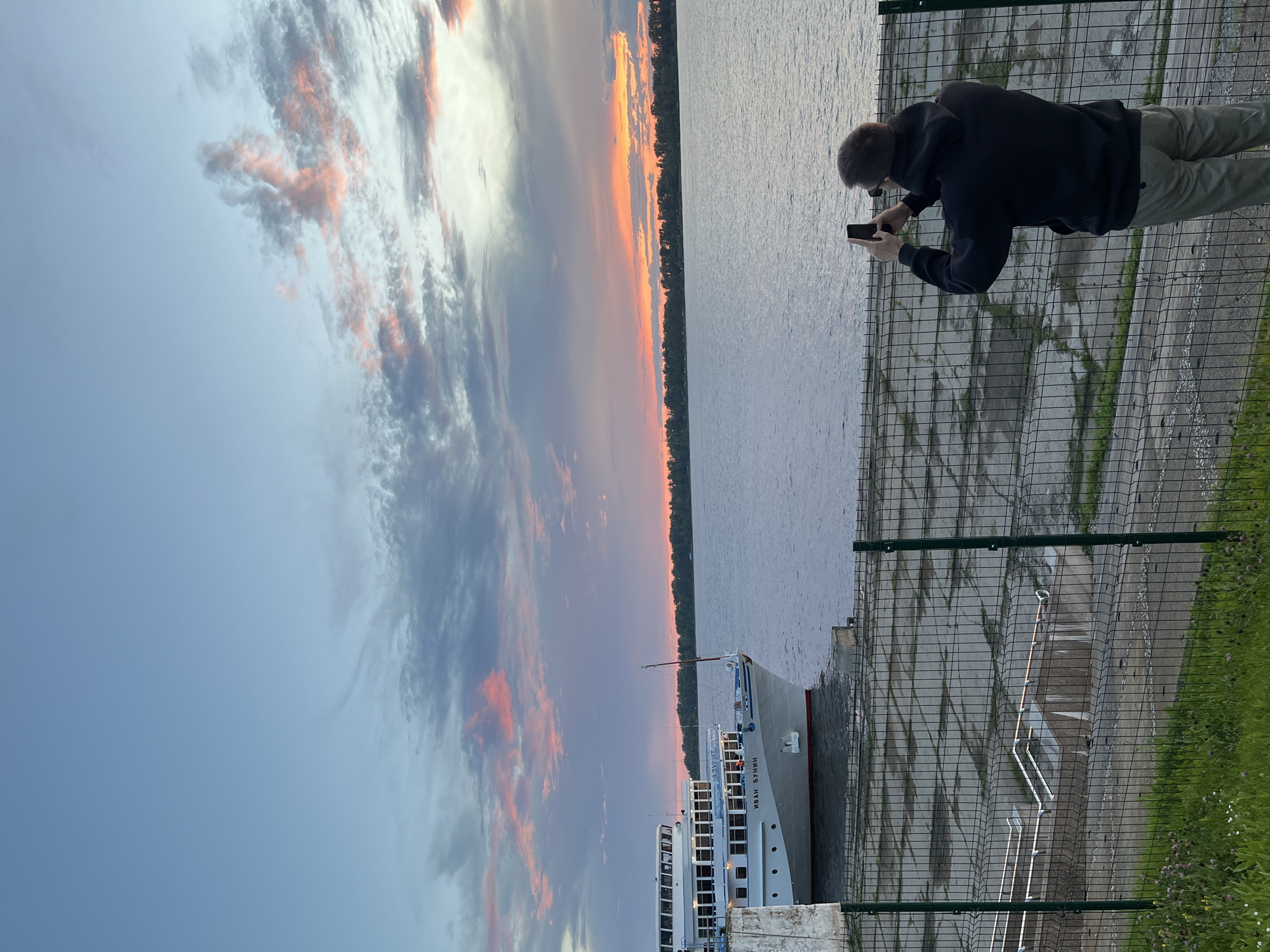The image depicts a person standing on a paved area near a body of water, possibly a river or lake, at sunset. The sky is filled with dramatic clouds painted in shades of orange, pink, and grey, indicating the end of the day. The water is calm and reflects the sky. A white ship with several decks is docked nearby, its name visible as "ИВАН БУНИН" in Cyrillic letters. The person is wearing a dark hoodie and light-colored pants, holding a smartphone and appearing to be taking a photo or video of the sunset. They are separated from the water by a wire fence. The ground is paved with concrete slabs, and there is a strip of green grass with some wildflowers to the right of the fence. The scene conveys a sense of peaceful observation of nature's beauty. The location is identified as Novokuybyshevsk, Russia.