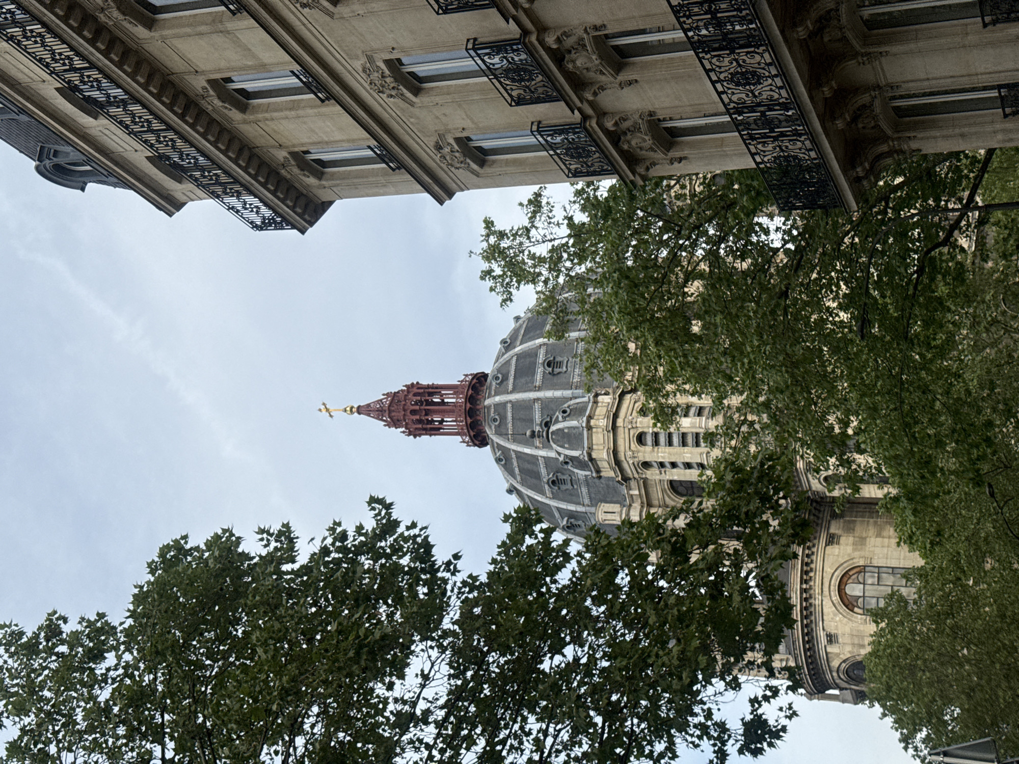 The image captures an upward-looking perspective of grand Parisian architecture, framed by lush green trees, under an overcast sky. In the mid-ground and right side of the frame, the magnificent dome of a building, likely a church, is the central focus. The dome features a dark, segmented roof, possibly of slate or metal, and is crowned by an elaborate reddish-brown spired lantern, topped with a golden cross. Ornate windows and architectural details adorn the dome's lower sections.
Across the upper portion of the image, a classic multi-story Haussmannian-style building in light-colored stone is visible. It showcases several windows with elegant black wrought-iron railings and decorative sculptural elements, indicative of Parisian residential or public buildings. The intricate patterns of the railings are clearly distinguishable.
Dense green foliage from several trees occupies the foreground and mid-ground, partially obscuring the base of the dome and the lower parts of the Haussmannian building. The leaves appear vibrant and full, suggesting a season of lush growth.
The sky is a uniform, bright grey, indicating an overcast or cloudy day, with no direct sunlight. The scene depicts a serene urban landscape devoid of people or specific activity, emphasizing the architectural beauty and natural elements. The architectural styles strongly suggest the setting is Paris, France. No visible text is present.