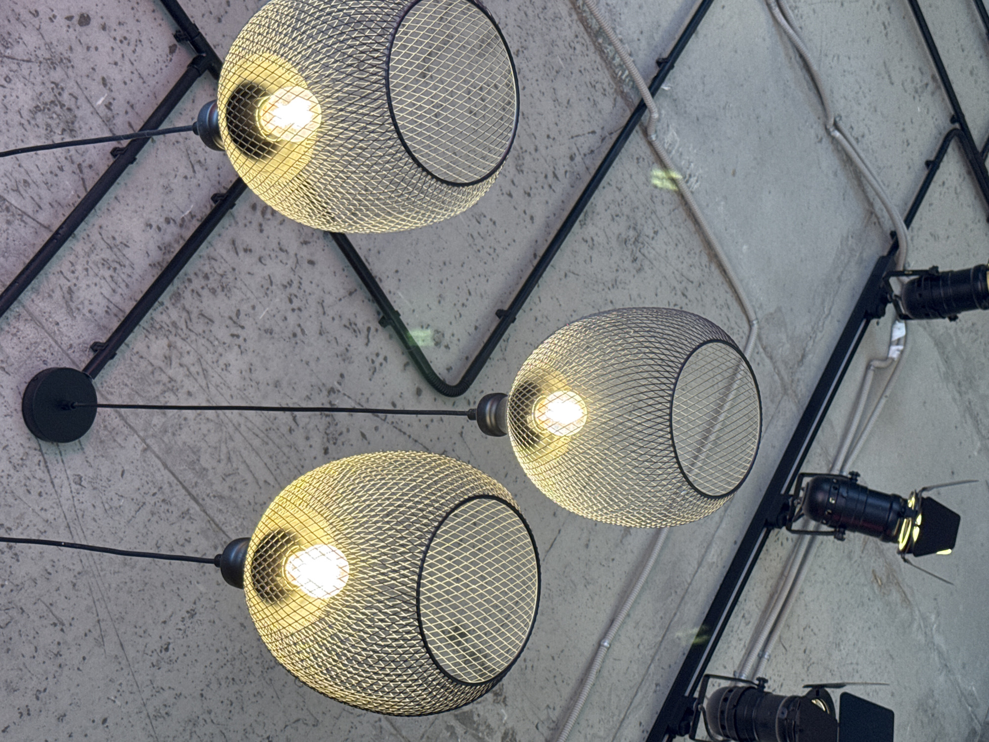 The image shows a close-up, low-angle view of a ceiling with industrial-style lighting fixtures. Three large, spherical lampshades made of wire mesh are hanging from the ceiling by black cords. Each lampshade encloses a lit lightbulb, casting a warm yellow glow. Below these, a black track lighting system is installed on the ceiling, from which several black cylindrical spotlights are suspended. The ceiling itself appears to be made of raw concrete with visible aggregate and has several gray conduits running across it. The overall aesthetic is modern and industrial. The image is likely taken indoors, possibly in a commercial space such as a restaurant, cafe, or studio, but no specific location in Saint Petersburg, Russia can be determined from the visual information. There are no people or animals visible in the image.