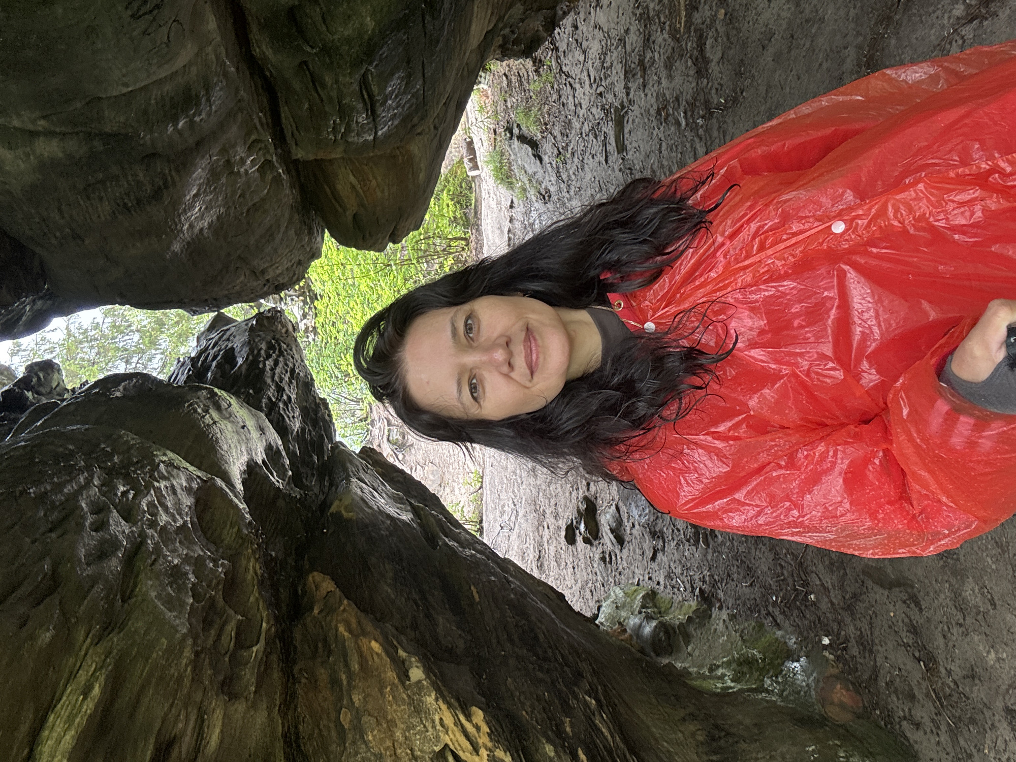 A medium shot captures a woman from the chest up, standing in a rocky alcove. She has dark, wavy hair and is looking towards the camera with a slight smile. She is wearing a bright red rain poncho over a dark garment. The alcove is formed by large, weathered rock formations, with the ground appearing to be a muddy path littered with small stones and twigs. Beyond the alcove, a sunlit forest with green foliage is visible. The image appears to be taken outdoors, likely in a natural park or trail. There is no information available to link this image to Shumikhinskii, Russia.