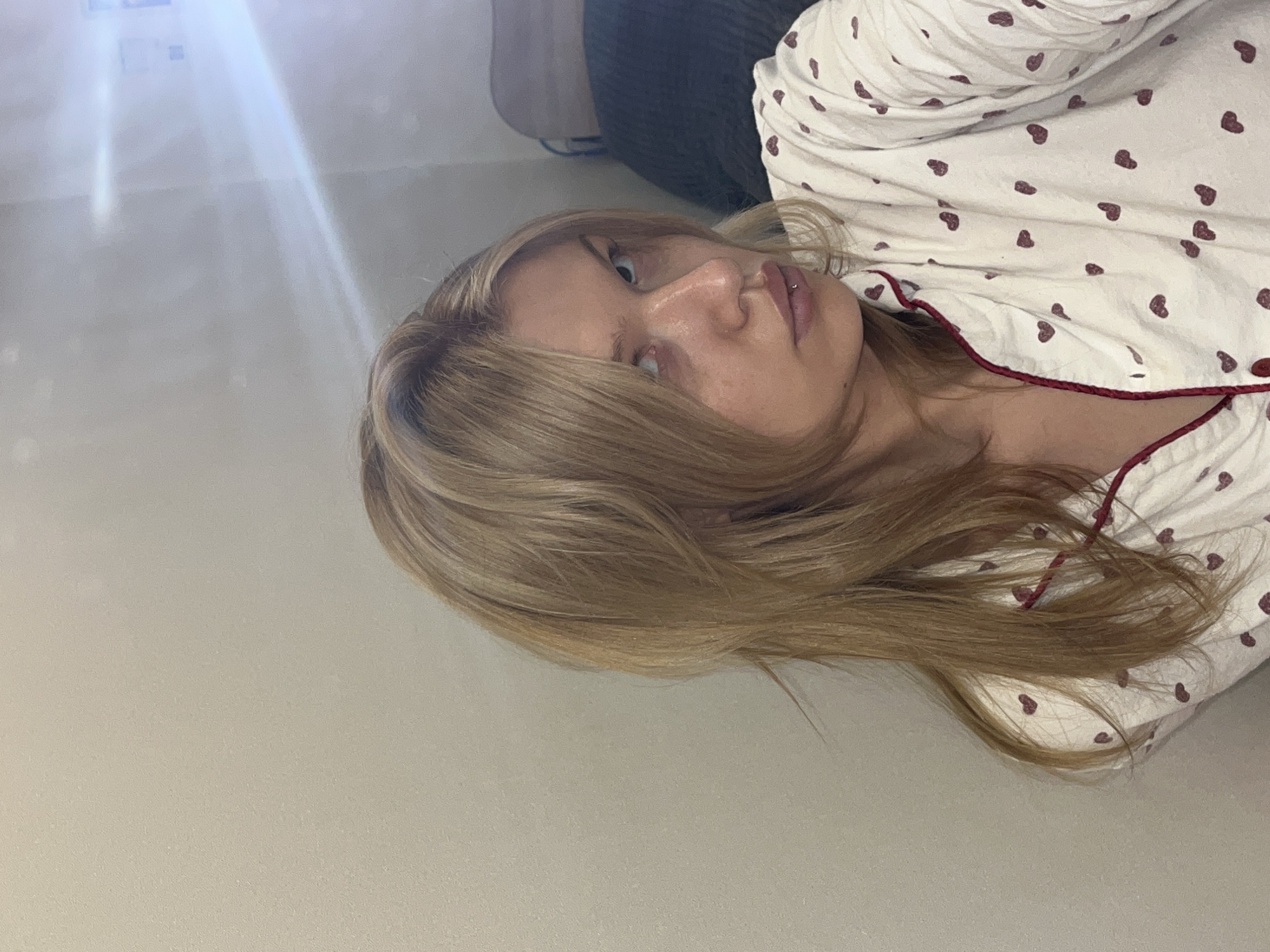 A blonde woman wearing a white top with red hearts is lying down. The background appears to be a light-colored wall. There is no visible evidence to suggest the location is Chaikovsky, Russia.