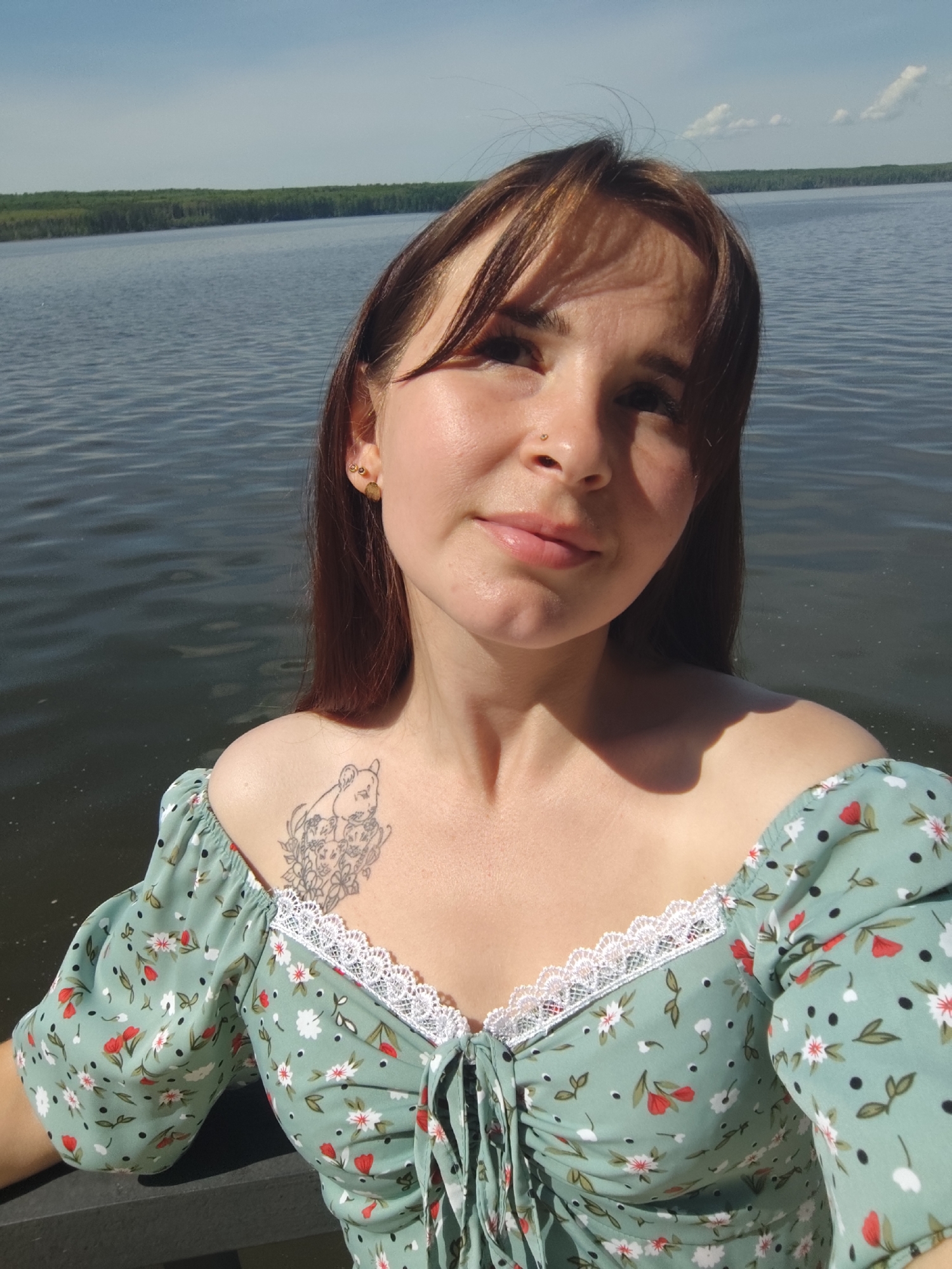 A woman is standing outdoors with a body of water behind her. The woman has brown hair with bangs, fair skin, and a nose piercing. She is wearing a green dress with white lace trim and a floral pattern. A tattoo is visible on her upper chest. The water behind her is calm, and trees line the distant shore. Based on information found online, the location may be in the city of Chaikovsky, Russia.