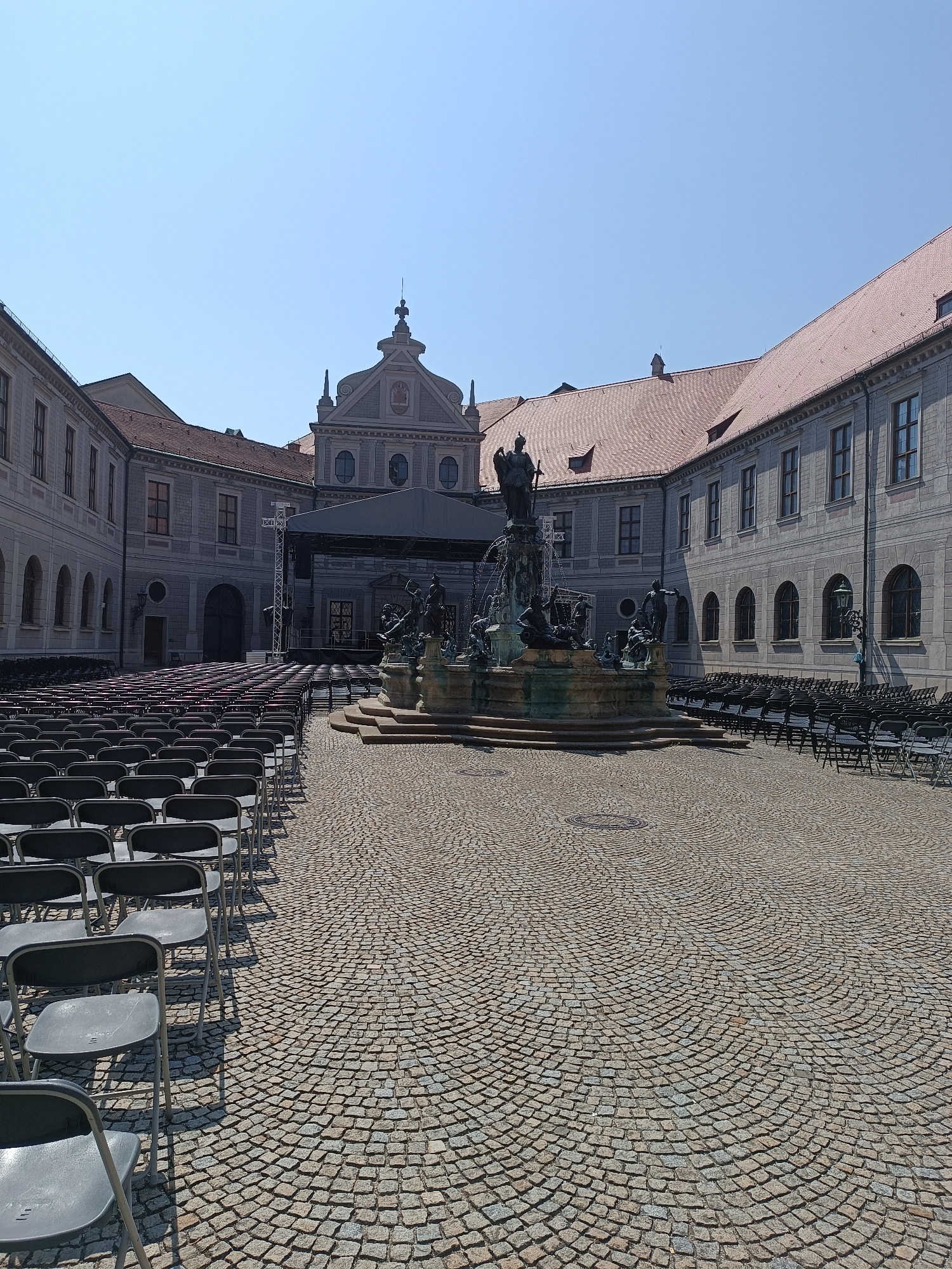 The image shows the courtyard of the Munich Residenz in Munich, Germany. The courtyard is paved with cobblestones and is set up with rows of black chairs, indicating it is prepared for an event. A stage with a canopy is visible in the background. In the center of the courtyard is a large, ornate fountain with several statues. The buildings surrounding the courtyard are made of stone with red tiled roofs, and the sky is clear and blue. There are no people visible in the image.