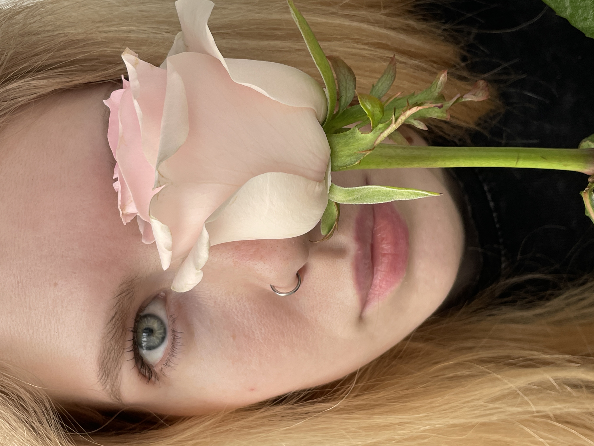 A close-up, rotated view depicts a fair-skinned individual with light blonde hair, lying down with a light pink rose superimposed over part of their face. The rose, in focus, obscures the upper portion of the person's face on the left side, covering the eyebrow and partially the eye. The stem of the rose extends across the bridge of the nose. A silver nose ring is visible. The background is primarily blonde hair, with a section of dark clothing on the upper right. The exact location in Izhevsk, Russia, cannot be determined from the visual information provided.