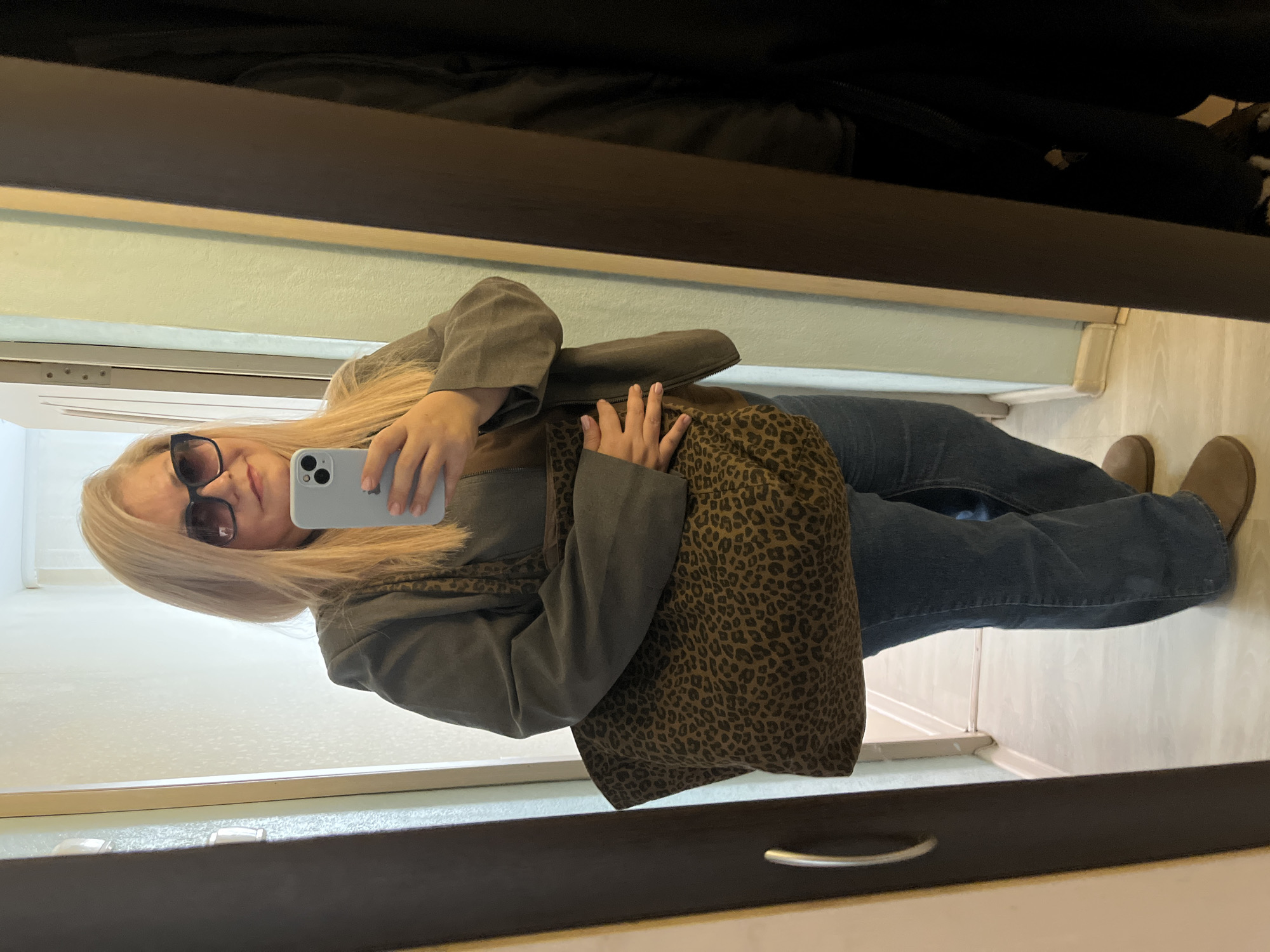 A woman takes a mirror selfie. She is wearing sunglasses, a green jacket, and blue jeans with brown shoes. She holds a light gray phone and is carrying a brown animal print bag. The location appears to be inside a home or apartment near a closet. There is no discernible indication of the city of Izhevsk, Russia.