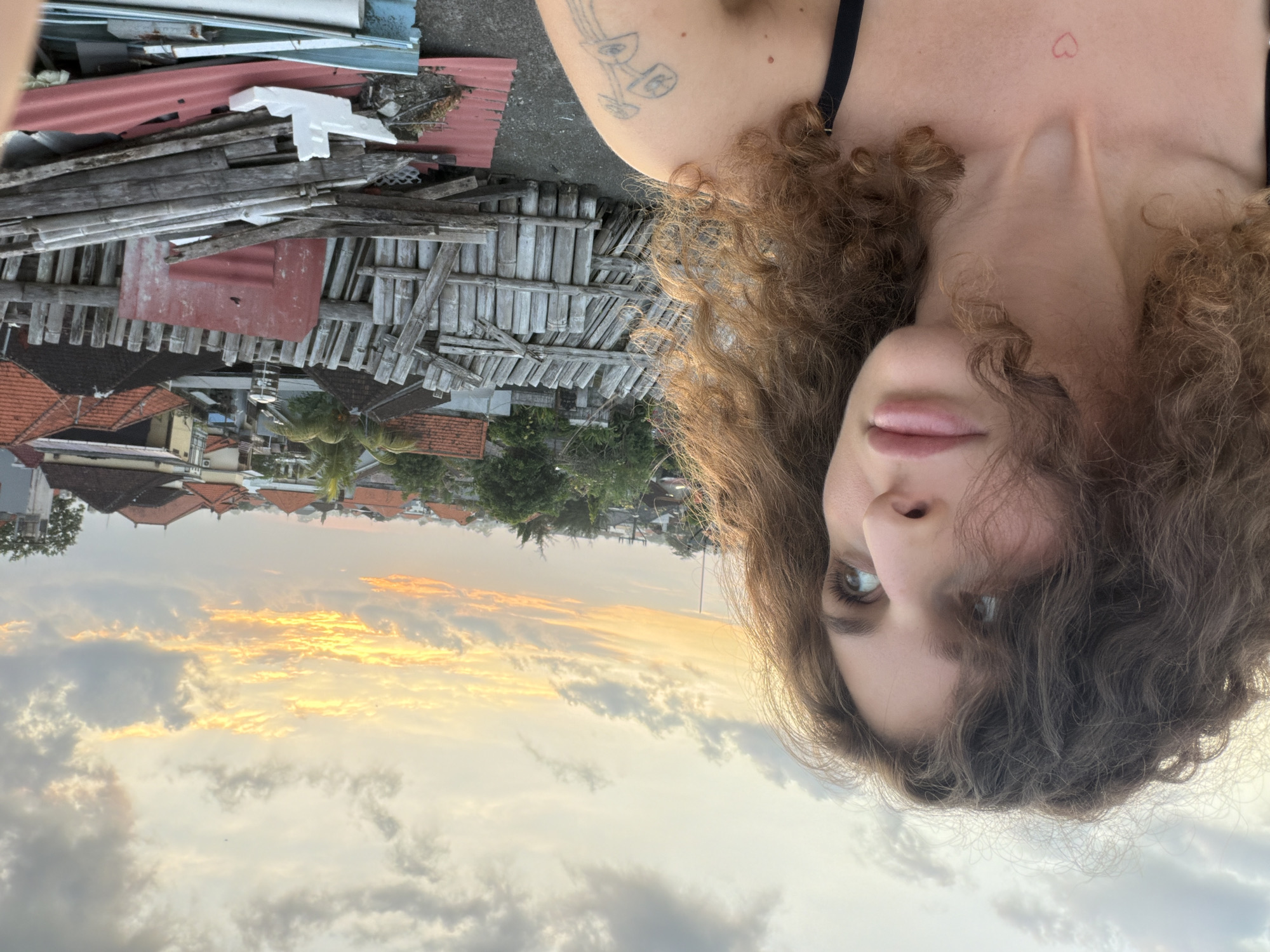 A medium shot captures a woman with curly brown hair looking to her right, with the sky and a city skyline in the background. The woman is positioned on the left side of the frame, facing away from the viewer. Her hair is voluminous and falls around her face and shoulders. A portion of her chest is visible, showing a small, red heart tattoo on her collarbone. She is wearing a black strap, suggesting she is wearing a tank top or bikini.
The background depicts a cityscape with buildings featuring red and brown tiled roofs. A makeshift fence constructed from weathered wooden planks and bamboo stalks runs horizontally across the mid-ground. Beyond the fence, trees and more buildings are visible, indicating an urban or semi-urban environment. The sky above is filled with clouds, with a portion of the setting sun casting a warm orange and yellow glow through the clouds. The lighting suggests it is either dawn or dusk. The scene appears to be from a rooftop in North Kuta, Indonesia, given the architectural style of the buildings and the lush vegetation. The overall impression is one of a casual moment captured outdoors, likely during sunset.