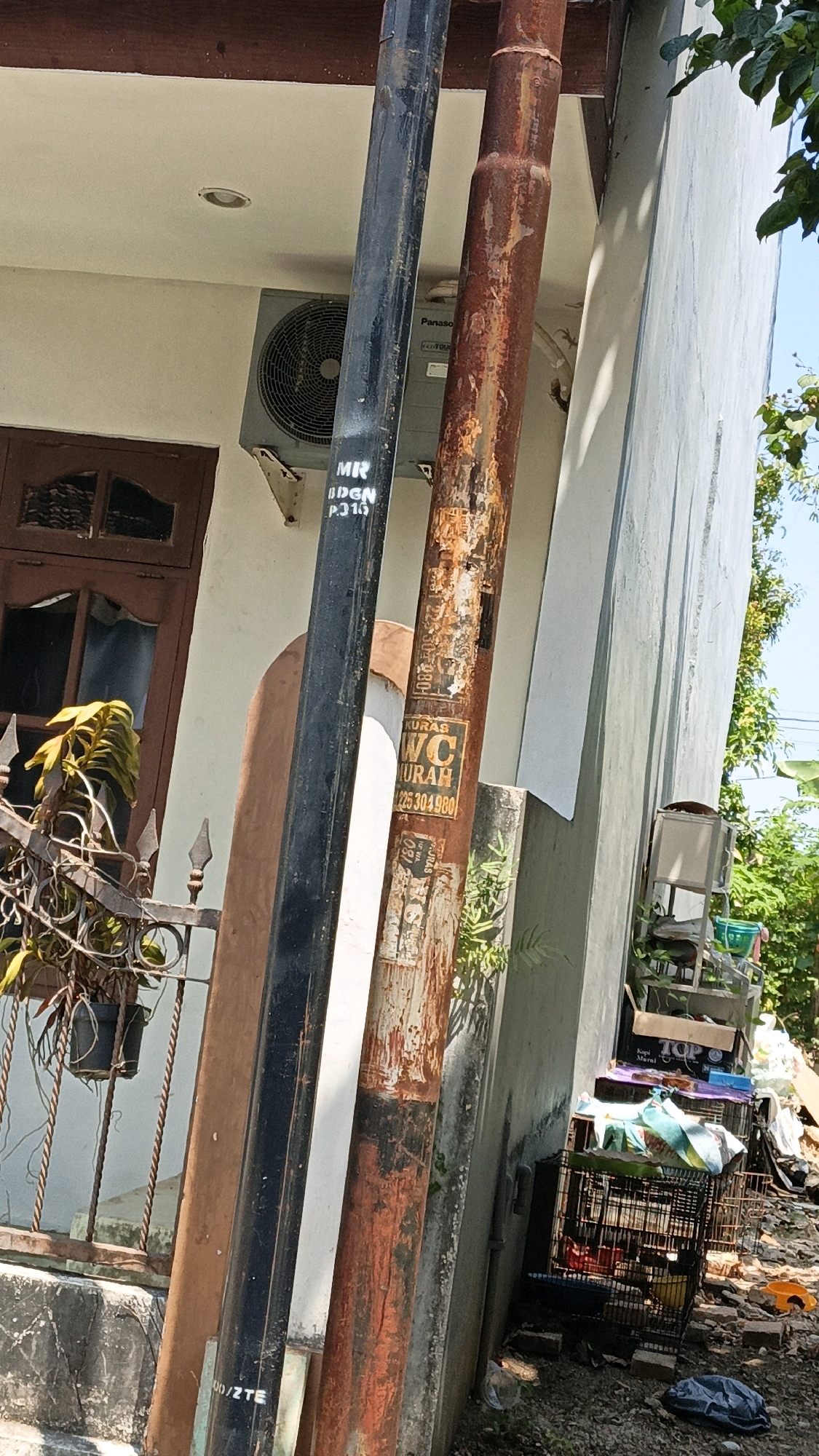 Two utility poles, one black and one rusty brown, are leaning against a light-colored building. The black pole has white lettering that appears to read "MR IDGN P.315". The rusty pole has several stickers, including one for "WC MURAH" with a phone number. An air conditioning unit is visible on the building wall. To the left of the poles, a dark brown wooden door with glass panes is partially visible, and a metal fence with a potted plant is in the foreground. To the right of the poles, more of the building is visible, along with some debris on the ground, including several cages, one of which contains birds. Trees and foliage are present in the background. The scene appears to be outdoors in a residential or commercial area.