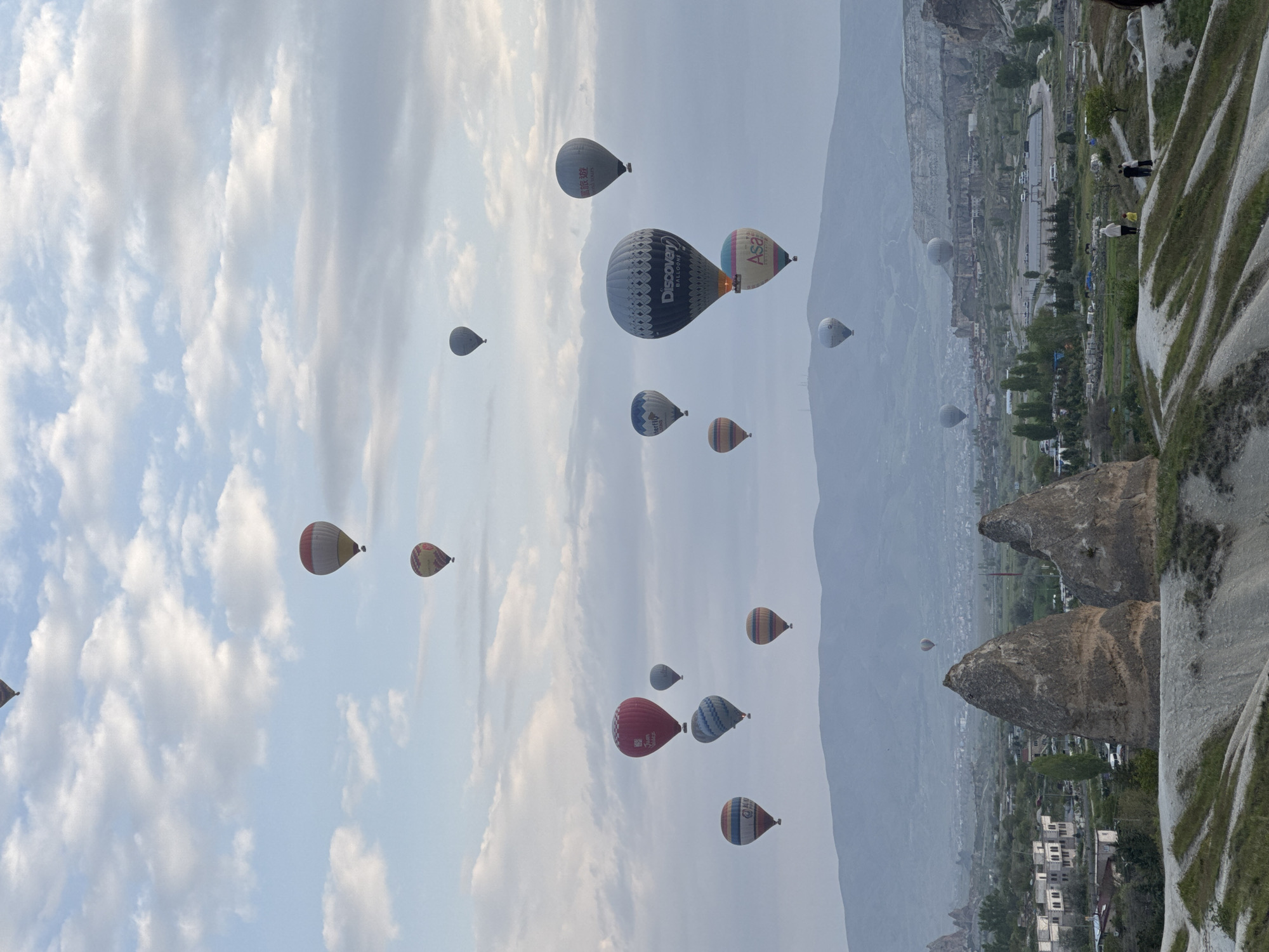 The image shows a sky filled with hot air balloons in Göreme, Turkey. The balloons are of various sizes and colors, some with company logos. In the background, there is a view of the landscape, featuring unique rock formations and buildings of the city, situated in a valley under a hazy sky. The horizon is indistinct due to the mist or haze.