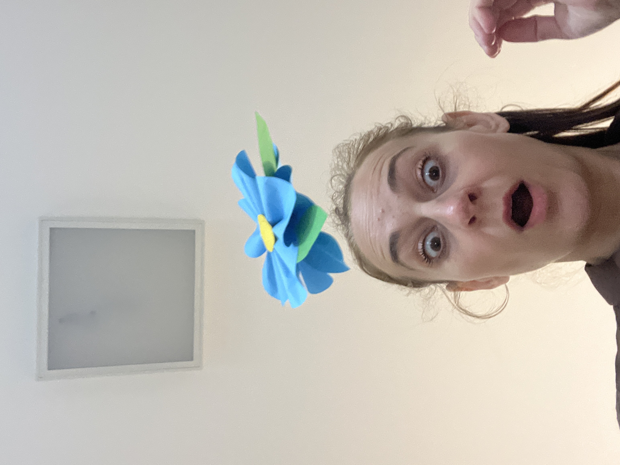 This is a vertically oriented, slightly blurry selfie of a young woman with wide eyes and an open mouth, suggesting surprise or shock. She is looking directly at the camera. Her hair is pulled back, and strands of lighter-colored hair frame her face. A hand with manicured fingernails is visible near the top of the frame.
In front of her, and slightly to the left, is a blue and green paper flower decoration, possibly part of a mobile or craft project. Behind the flower and to the left, a square, white, framed object with a frosted or translucent surface is visible, possibly a light fixture or a vent.
The background is a plain, light-colored wall or ceiling. The lighting appears to be indoor and diffuse. The overall impression is of a spontaneous, perhaps playful, moment captured indoors. There is no visible text in the image.