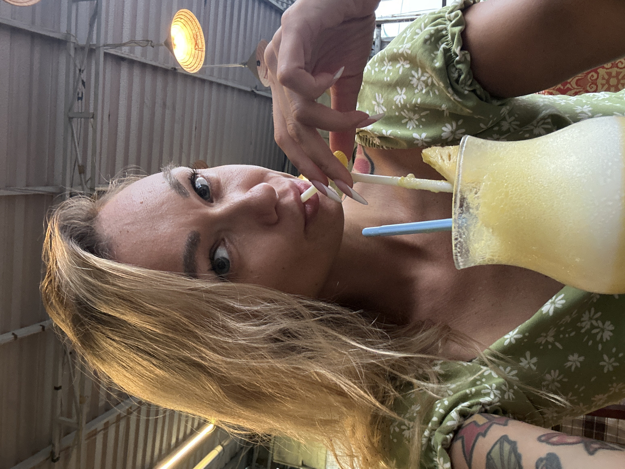 A woman is holding a yellow pineapple juice drink in Phu Thuy Ward, Vietnam. The woman has blonde hair, fair skin, and a green floral dress. She has long white nails and is holding a white straw in her mouth. The drink has a pineapple garnish on the rim. The background appears to be a building with corrugated metal siding and overhead lighting.