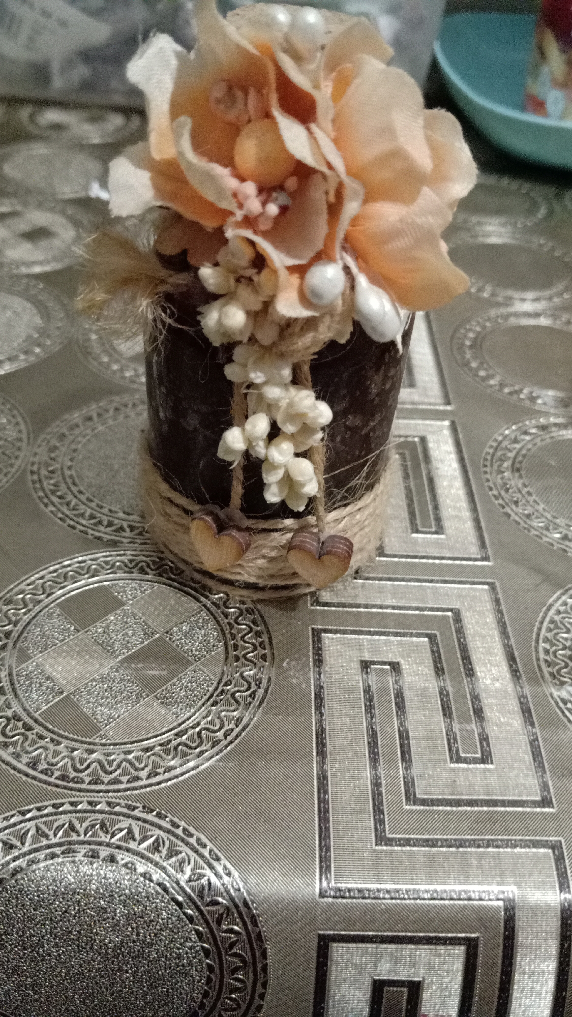A still life features a decorative floral arrangement in a dark brown cylindrical container. The arrangement includes peach-colored fabric flowers with smaller white pearl-like accents and a few sprigs of a dry, straw-like material. The container is adorned with natural twine wrapped around its upper and lower sections. Two small wooden heart ornaments hang from the twine.
The arrangement sits on a silver tablecloth with a repeating pattern of concentric circles and geometric designs. A blurry turquoise object is visible in the upper right corner of the image.
The image does not provide sufficient details to determine the location in the city of Yerevan, Armenia.